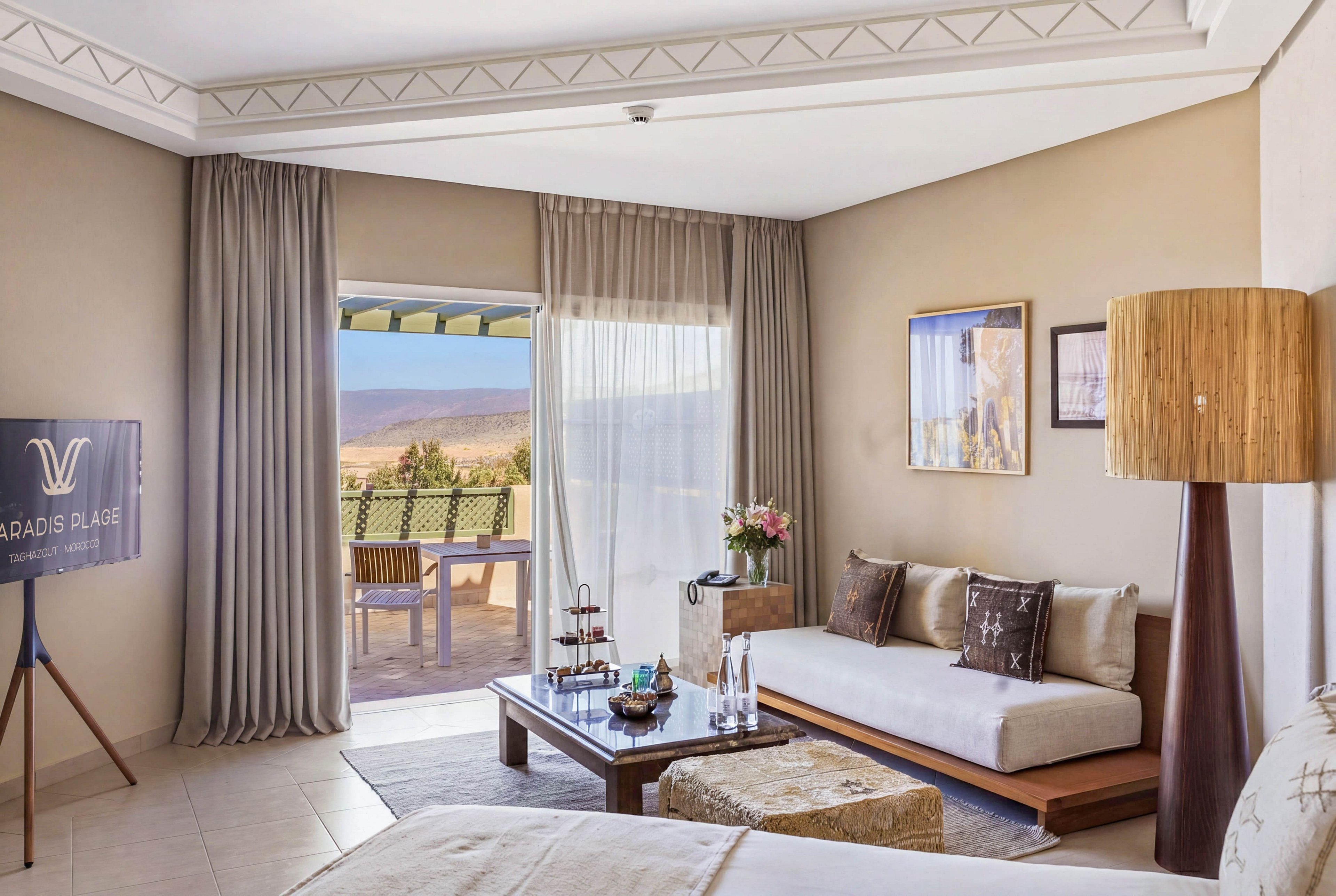A modern hotel suite features a neatly made bed and a cozy seating area with a low sofa and cushioned ottomans. Large curtains frame a sliding glass door that opens onto a terrace with a table and chairs overlooking a scenic coastal landscape. A wooden floor lamp, decorative pillows, and a coffee table with water bottles and flowers add warmth and comfort to the room. A display screen on the left shows the text PARADIS PLAGE with a stylized emblem.