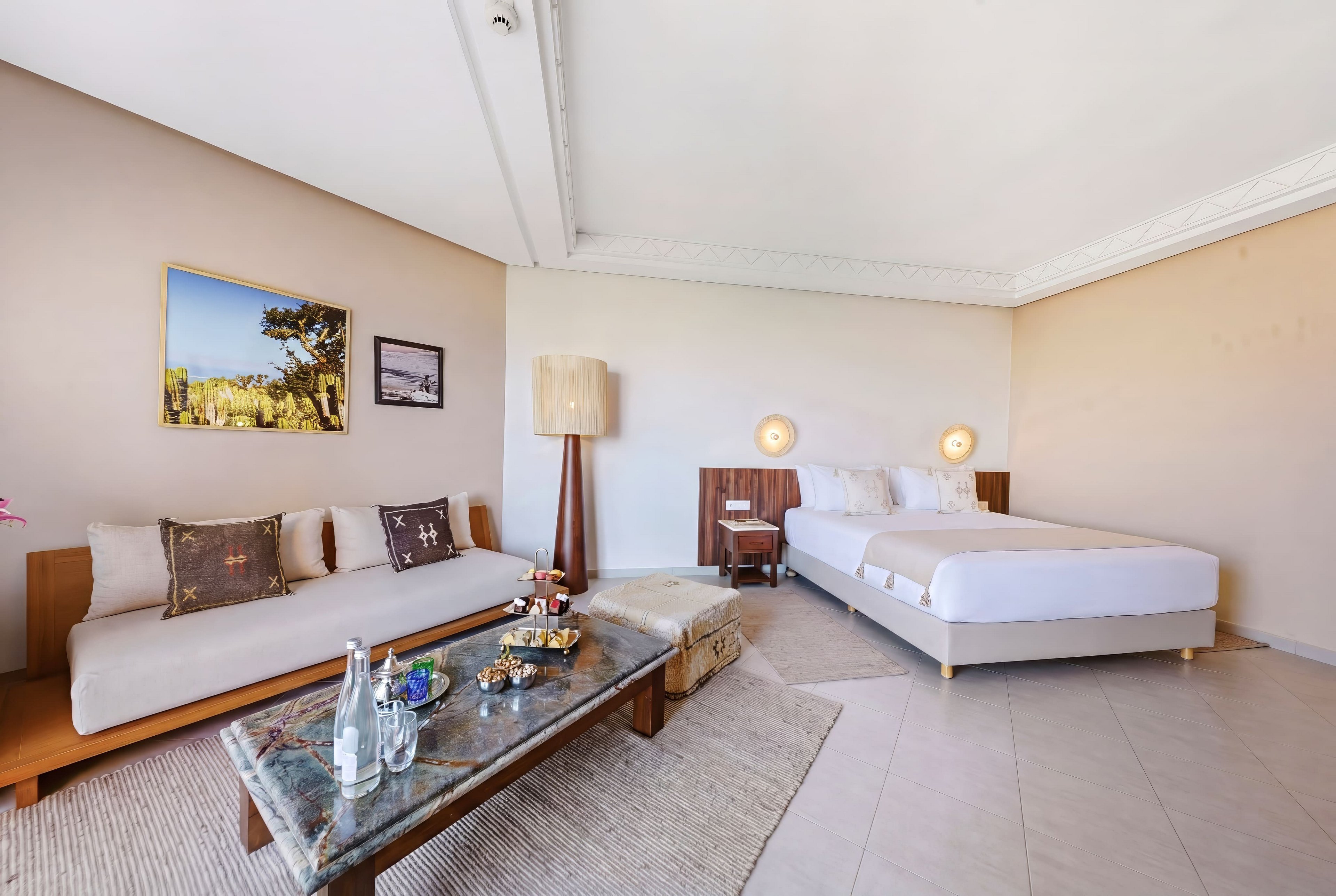 A spacious modern hotel suite features a large bed and a minimalist lounge area. A low sofa with decorative pillows faces a stone coffee table set with drinks and snacks. Neutral walls, light flooring, and a large floor lamp create an airy, contemporary atmosphere. Framed artwork on the wall and soft lighting add a relaxed, welcoming feel to the room.