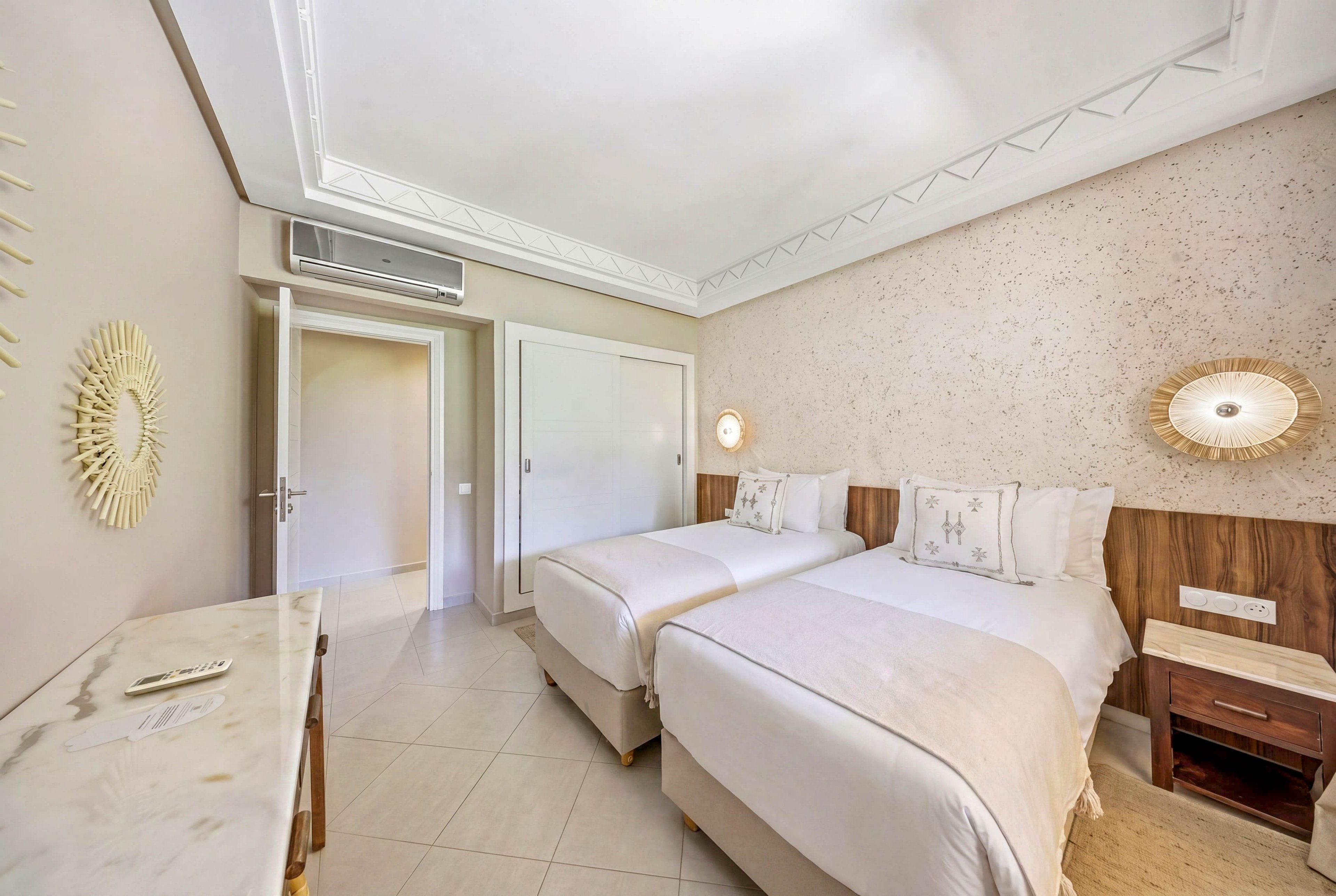 A bright, modern hotel room features two neatly made twin beds with white linens and light beige accents. The space includes a wooden nightstand, wall-mounted lamps, and a decorative mirror on the left wall. A marble-topped console table and built-in wardrobe add to the clean, minimalist design. Soft neutral tones and ample lighting create a calm, inviting atmosphere.