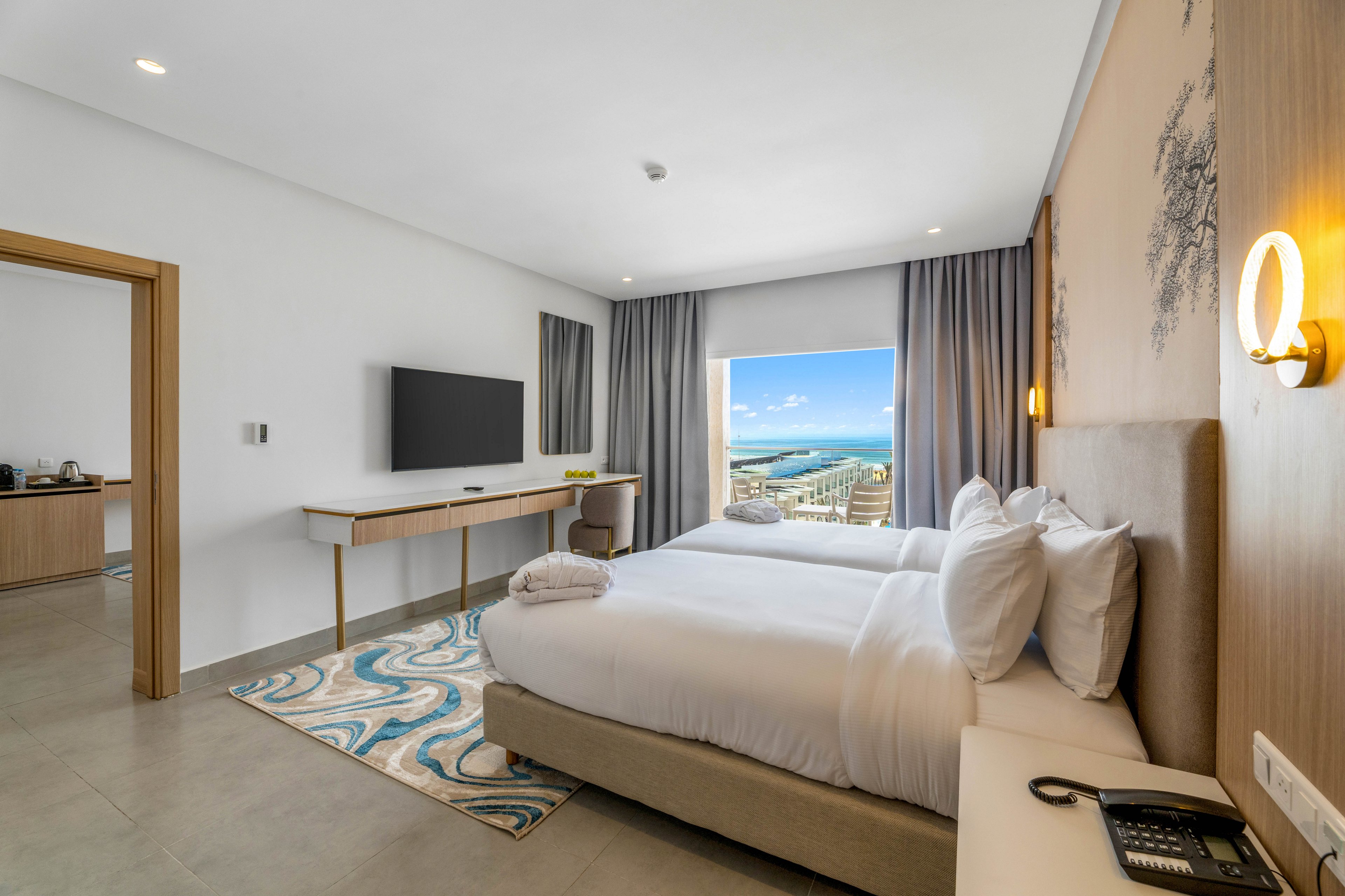 A contemporary hotel bedroom features two neatly made single beds with white linens and plush pillows. The room includes a wall-mounted flat-screen TV, a minimalist wooden desk, and a patterned rug adding color to the neutral decor. Large curtains frame a window that reveals a bright oceanfront view with blue water and a coastal promenade. An adjoining room with a small refreshment area is visible through an open doorway, enhancing the sense of space and comfort.