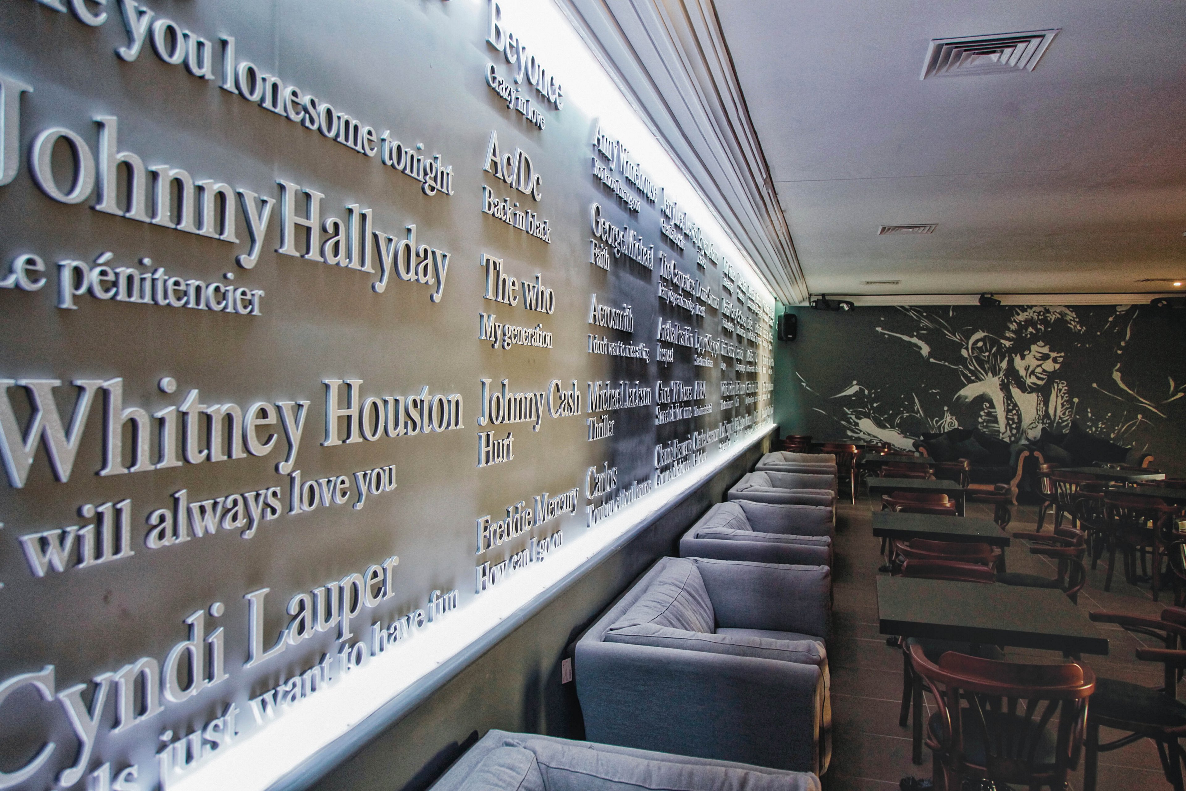 The main subject is a stylish lounge featuring a wall decorated with illuminated names and song titles of famous music artists such as Johnny Hallyday, Whitney Houston, and Cyndi Lauper. The setting includes comfortable gray armchairs and wooden tables, creating a relaxed and contemporary atmosphere. The visuals highlight the prominent text on the wall, soft lighting, and a monochrome mural in the background. No explicit numeric values are visible in the image.