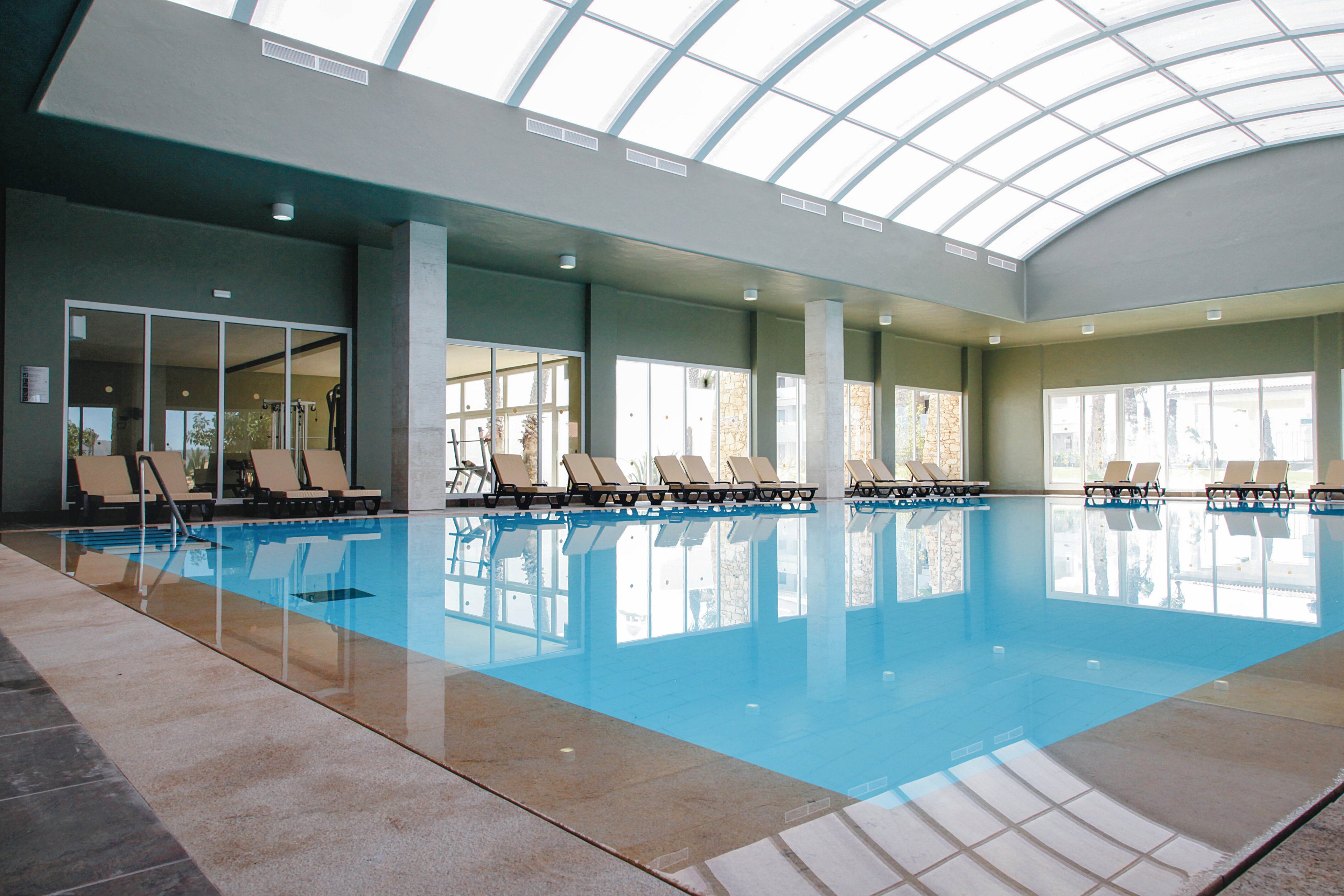 A spacious indoor swimming pool is featured in a contemporary setting with a high, curved glass ceiling. Numerous lounge chairs are arranged along the poolside, and large windows allow natural light to fill the area. The clean lines and neutral color palette create a calm and inviting atmosphere, ideal for relaxation or leisure swimming.