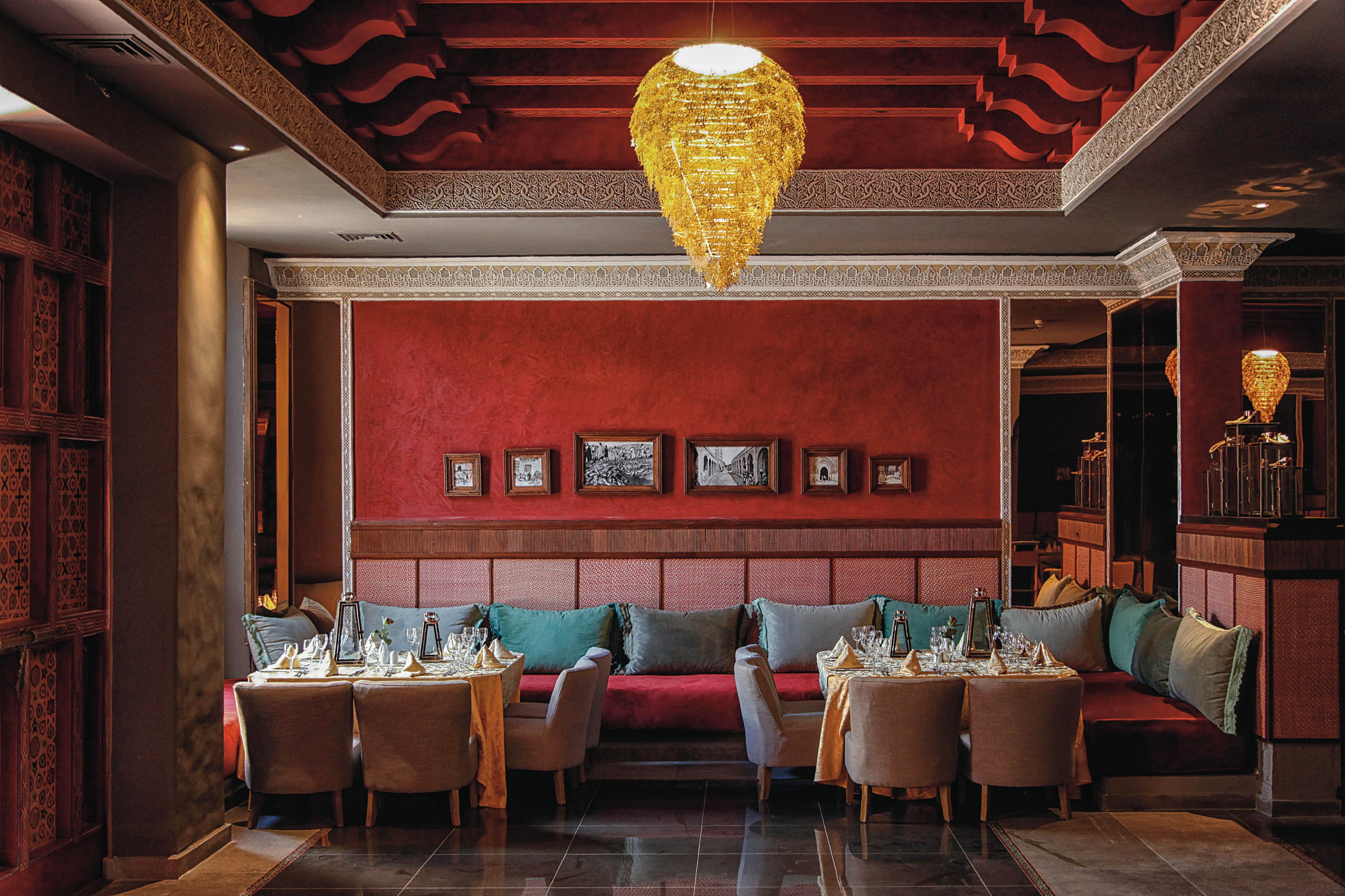 The image features a sophisticated restaurant setting with plush seating and tables set for dining. A striking golden chandelier hangs from the ceiling, illuminating the rich red walls adorned with framed artwork. The mood is warm and inviting, with a palette of deep reds, golds, and neutral tones. The scene is empty of people, focusing on the ambiance and decor.