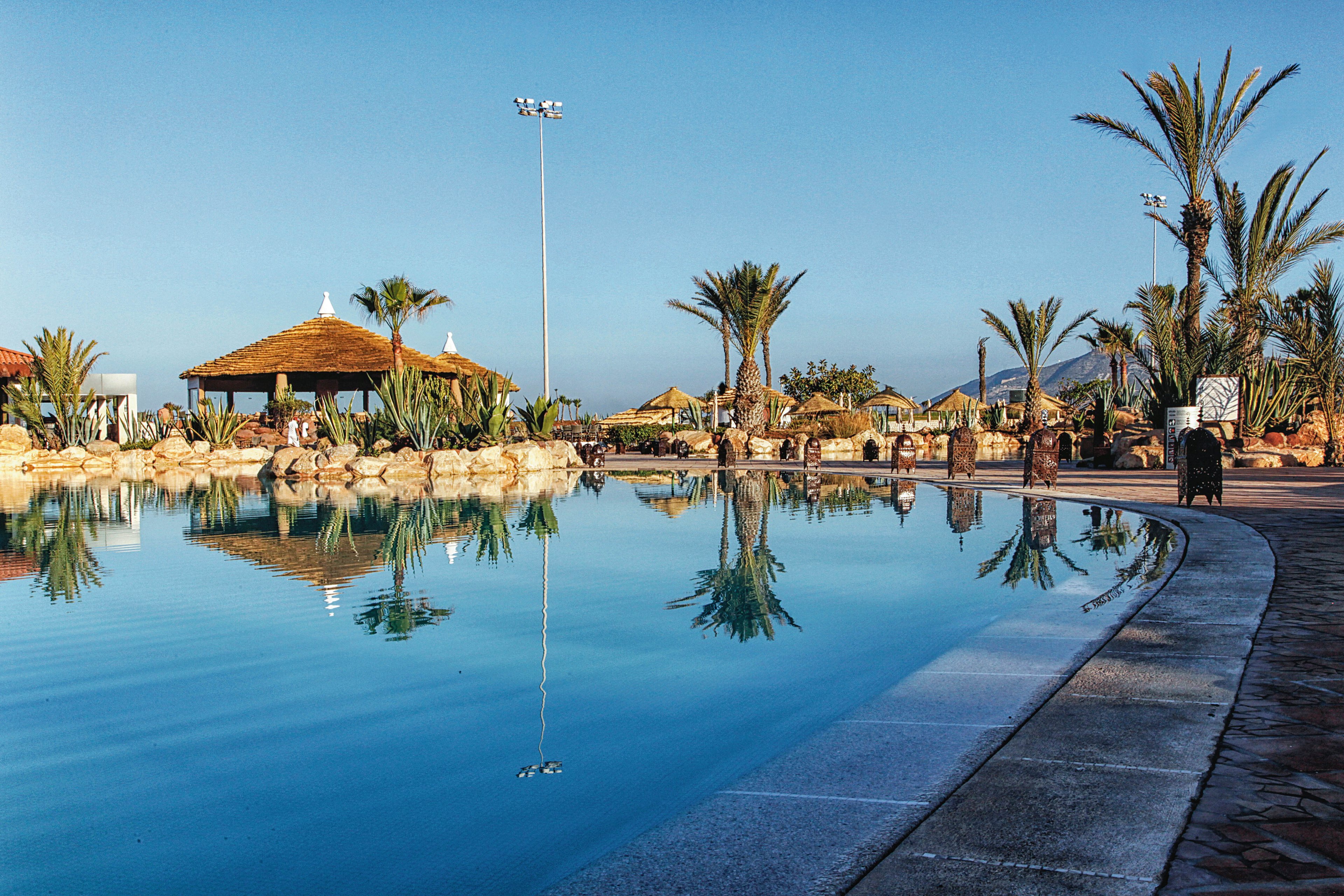 A large outdoor swimming pool is surrounded by palm trees and thatched-roof cabanas, creating a tropical resort atmosphere. The scene is set under clear blue skies with mountains visible in the background. The water reflects the surrounding landscape, and the area appears calm and inviting, ideal for relaxation. No visible text or numeric values are present in the image.