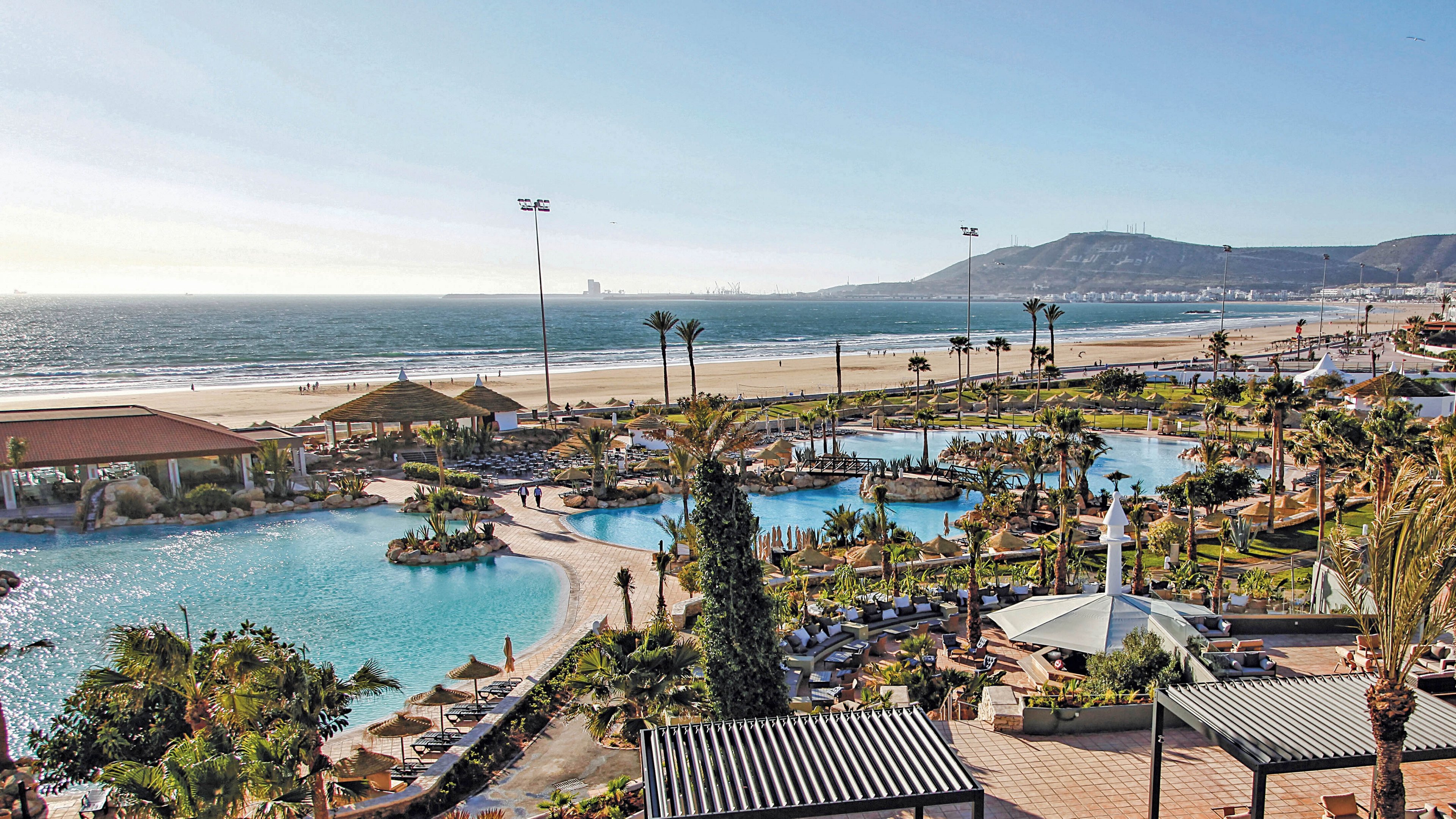 A sprawling resort complex overlooks a sandy beach and the ocean, featuring multiple swimming pools surrounded by palm trees and sun loungers. The setting is lively and sunlit, with guests relaxing under shaded cabanas and walking along tiled pathways. The mood is vibrant and leisurely, with a clear blue sky and distant hills enhancing the scenic coastal atmosphere.