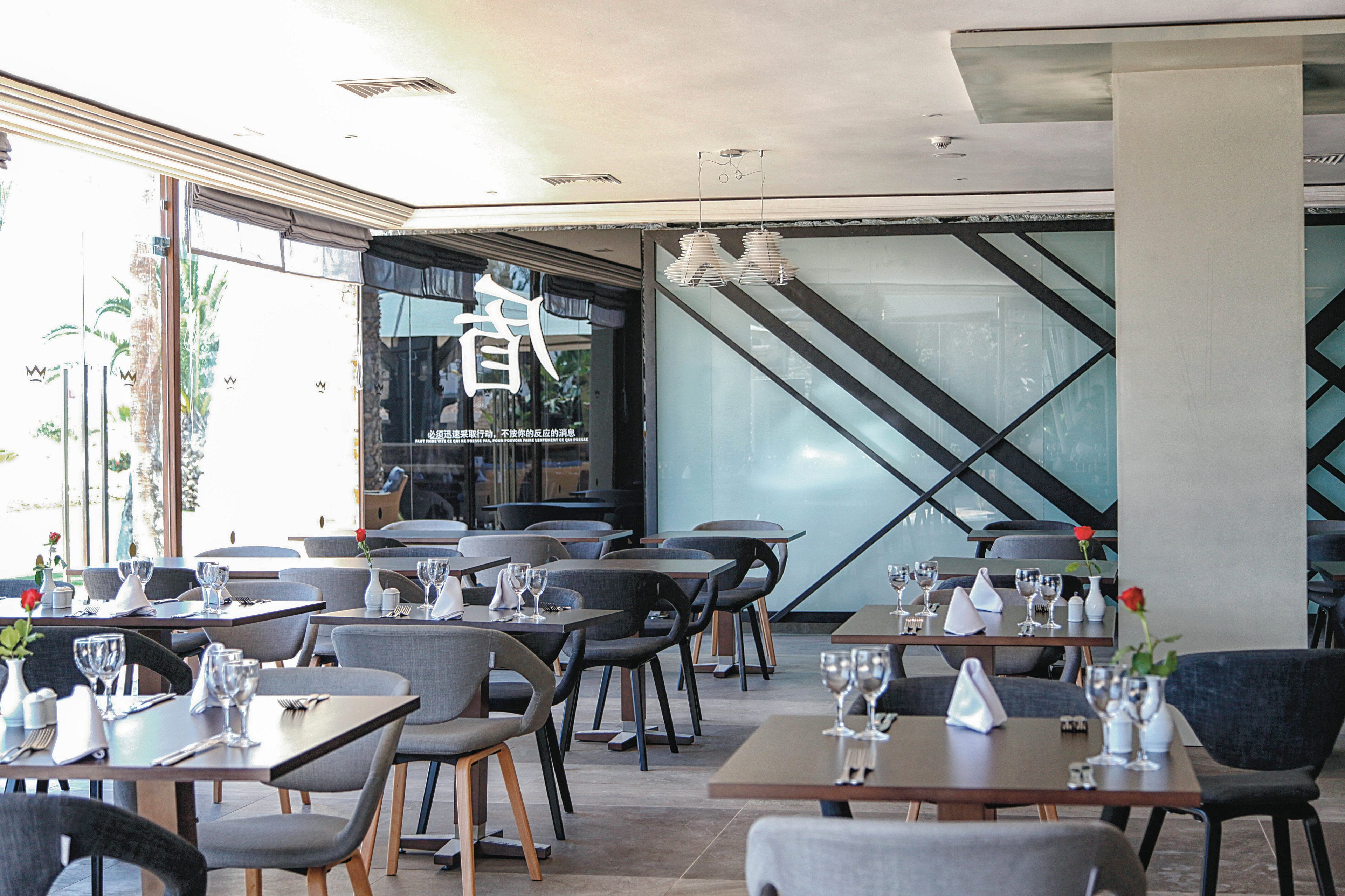 A contemporary restaurant dining area features neatly arranged tables with glassware, napkins, and single red roses. The space is well-lit with natural light from large windows and has a minimalist design with geometric glass wall accents. A visible Asian character is displayed on the glass, adding a unique visual element. The overall atmosphere is clean, inviting, and ready for guests.