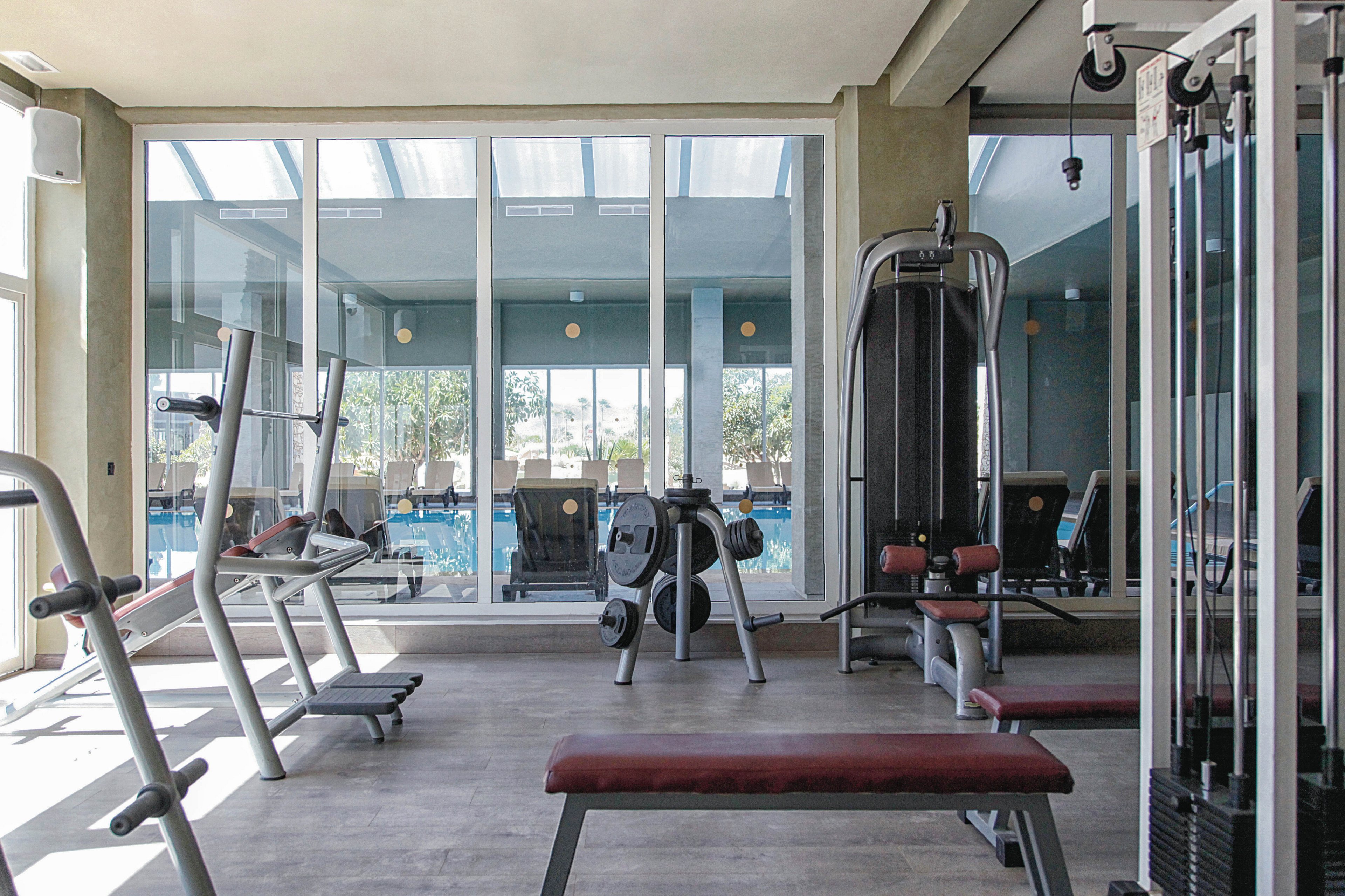 A contemporary fitness room features various exercise machines, including treadmills, stationary bikes, and weight training stations. The gym is well-lit with natural light streaming through large windows, offering a view of an outdoor pool area. The space is clean and organized, with muted tones and sleek design elements. No visible text or numeric values are present on the equipment.