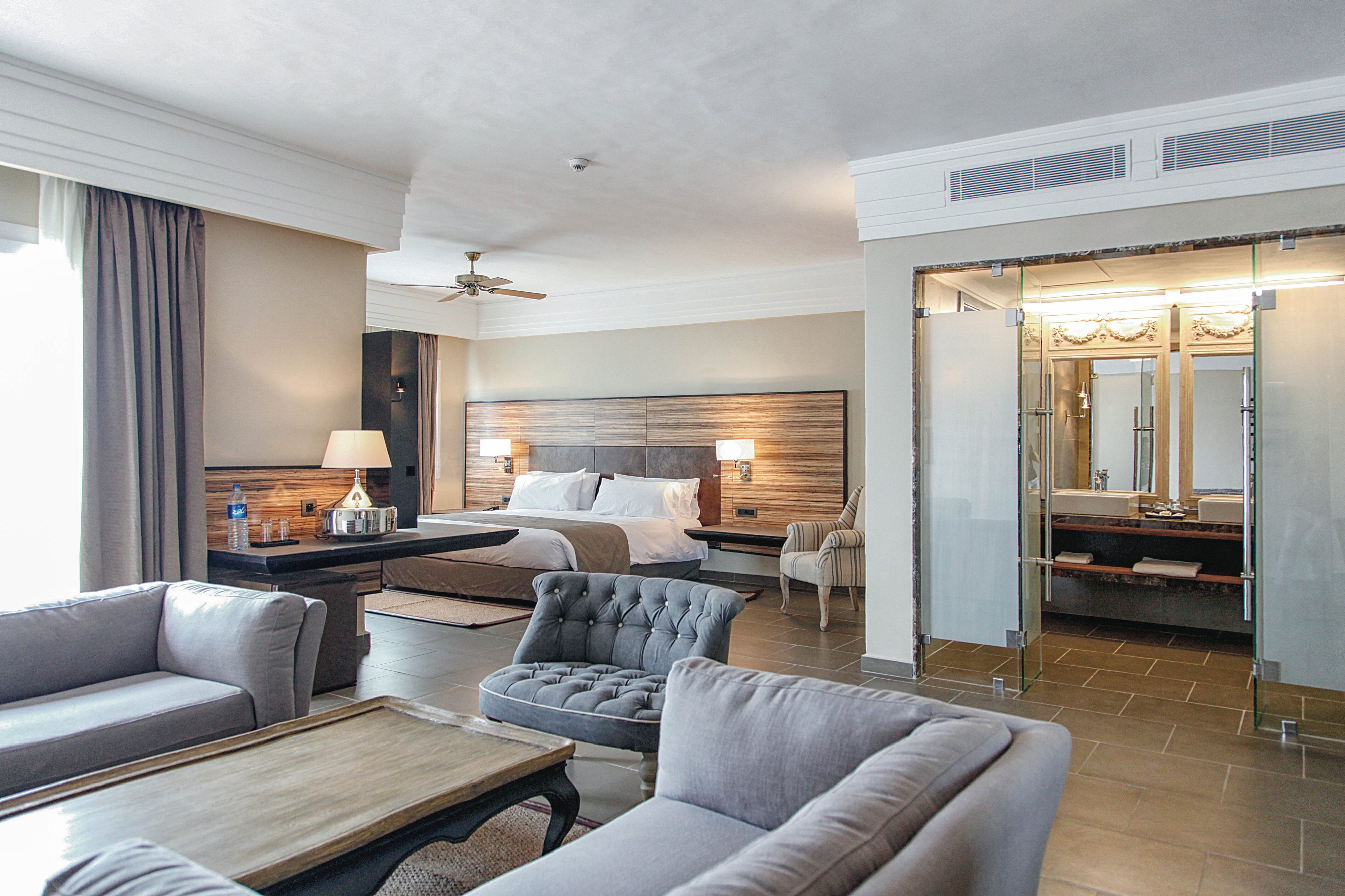 A contemporary hotel suite features a large bed, stylish wooden headboard, and a comfortable seating area with plush sofas and a coffee table. The room is well-lit with natural light from a large window and includes a glass-walled bathroom visible in the background. Neutral tones and elegant decor create a relaxing and inviting atmosphere.