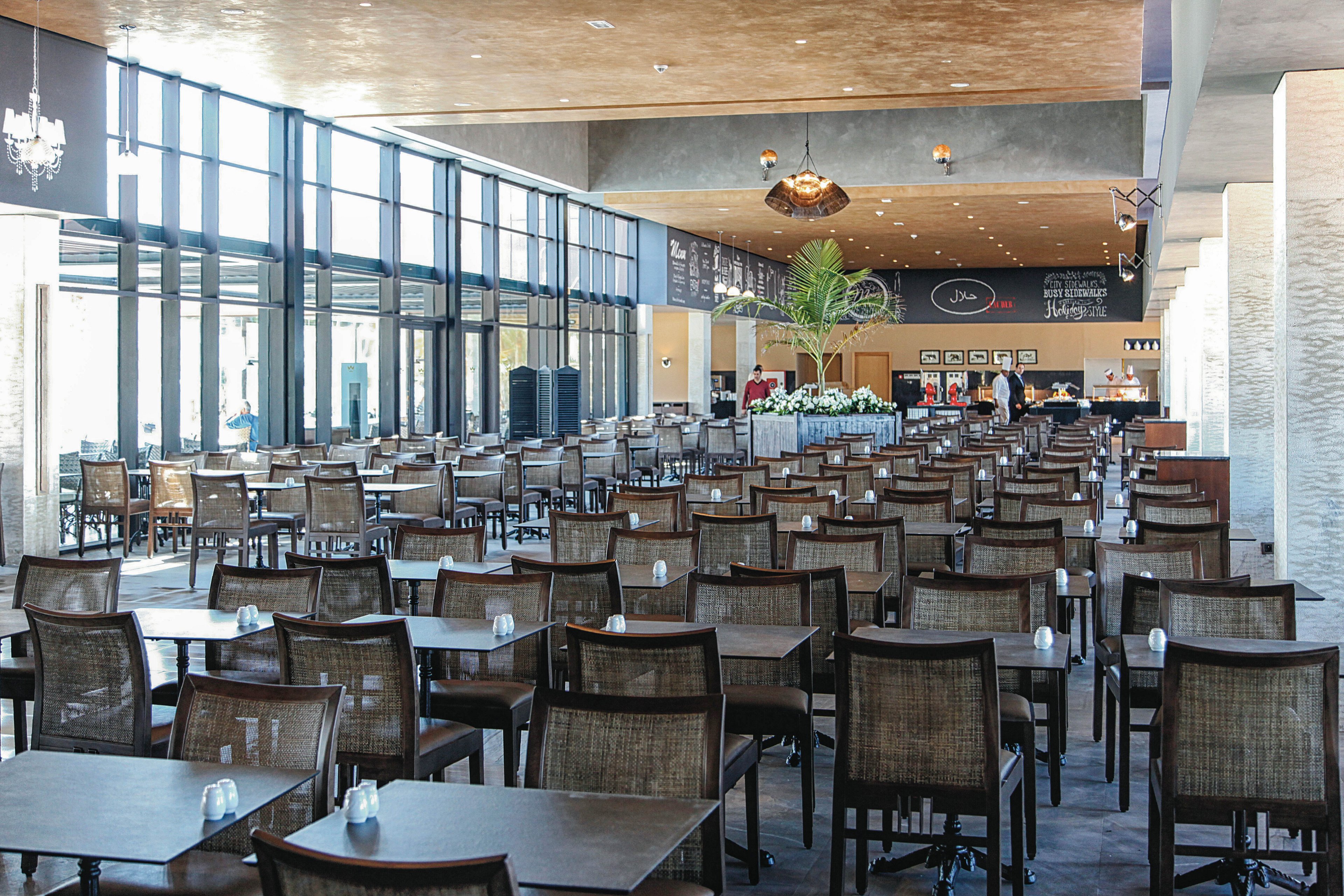 A large, contemporary restaurant dining area features rows of empty wooden tables and chairs arranged neatly. The space is filled with natural light from floor-to-ceiling windows on one side, and a decorative ceiling with modern lighting fixtures. A counter area and chalkboard menu are visible in the background, along with a prominent indoor plant adding greenery to the setting.
