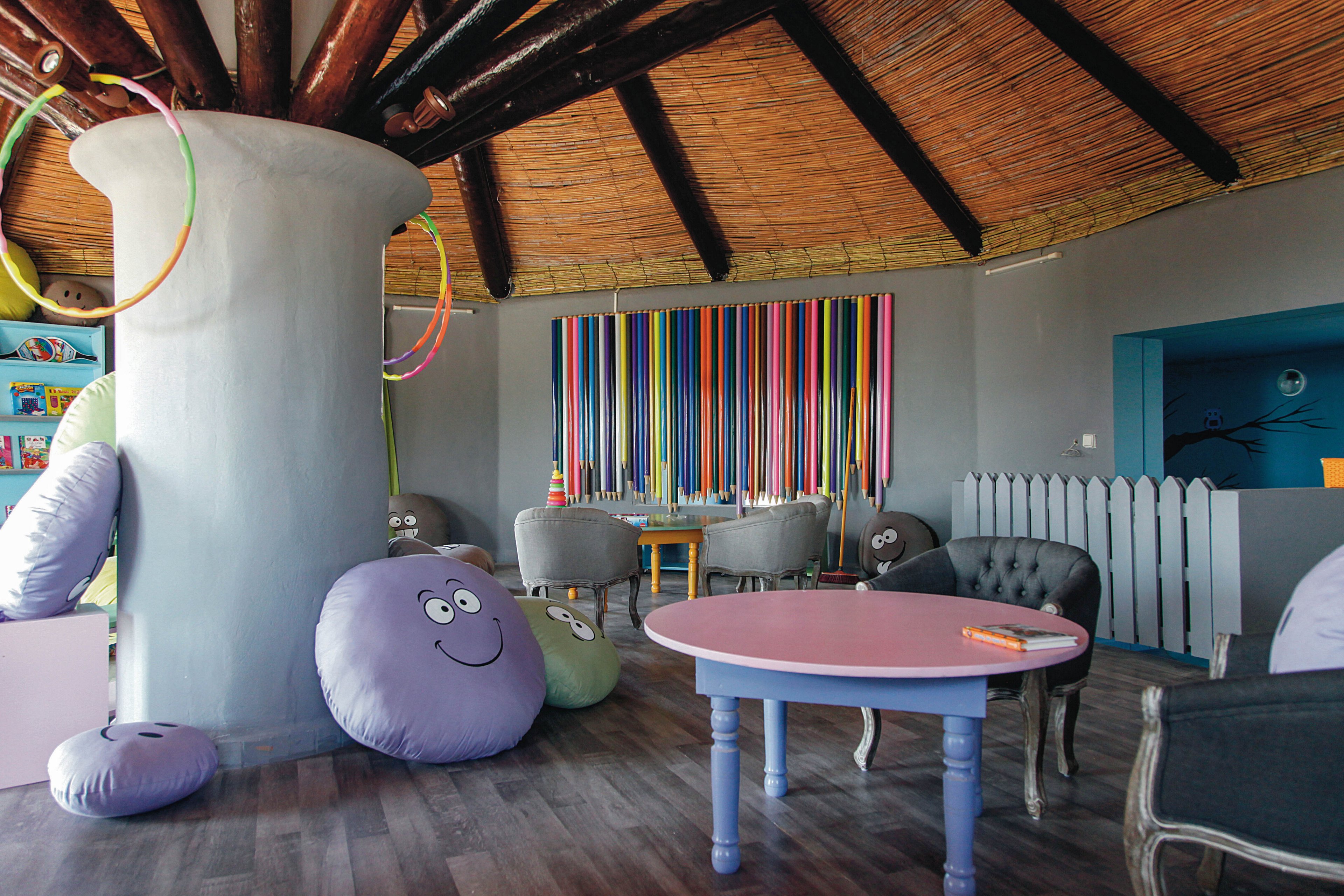 The image shows a vibrant indoor playroom featuring large, round cushions with smiling faces, a pastel-colored table, and whimsical decor. The setting includes a wooden ceiling, a central pillar, and colorful vertical stripes on the wall. The room is furnished with soft seating and playful elements, creating a cheerful and inviting atmosphere for children. No visible numeric values or identifiable brands are present.