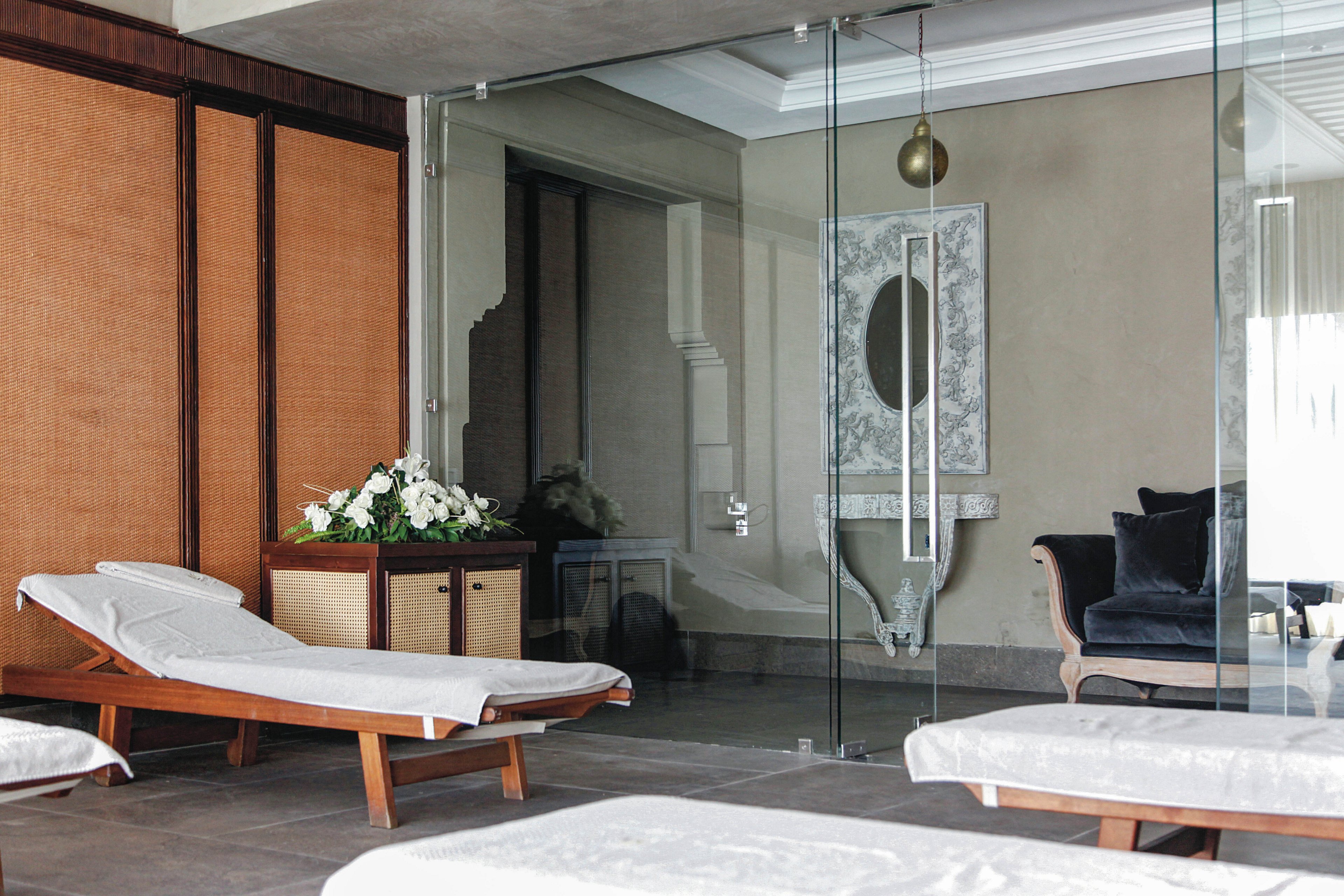 A tranquil spa relaxation room features several cushioned lounge chairs arranged for comfort. The setting includes a glass wall, neutral tones, and a decorative console with a floral arrangement. The space is well-lit, with contemporary furnishings and a calm, inviting atmosphere.