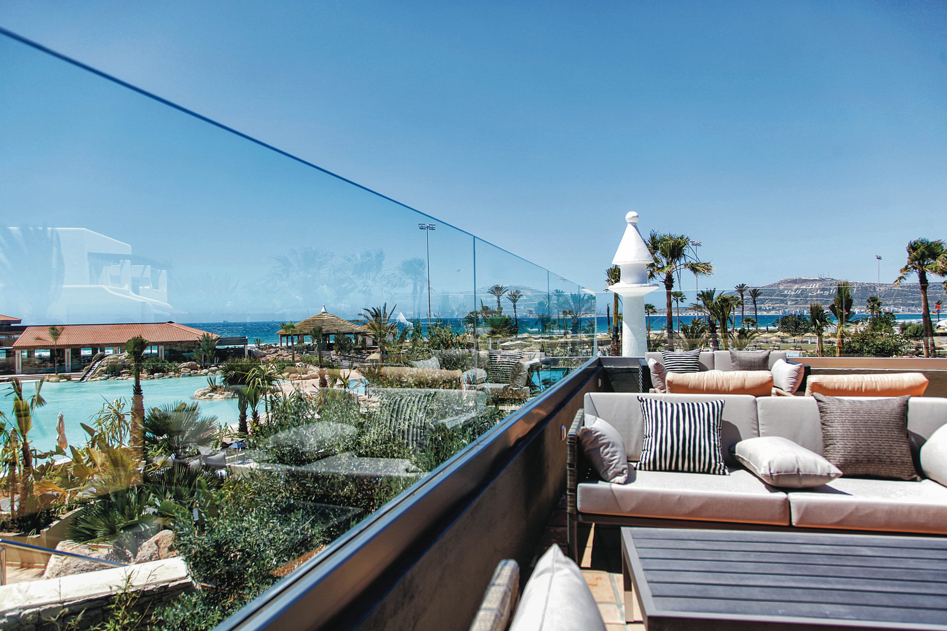 A modern outdoor seating area with plush cushions is set on a terrace, separated by a glass railing. The view overlooks a resort pool surrounded by palm trees, with the ocean and distant hills visible under a clear blue sky. The scene is bright and inviting, featuring neutral and earth-toned decor with striped accent pillows.