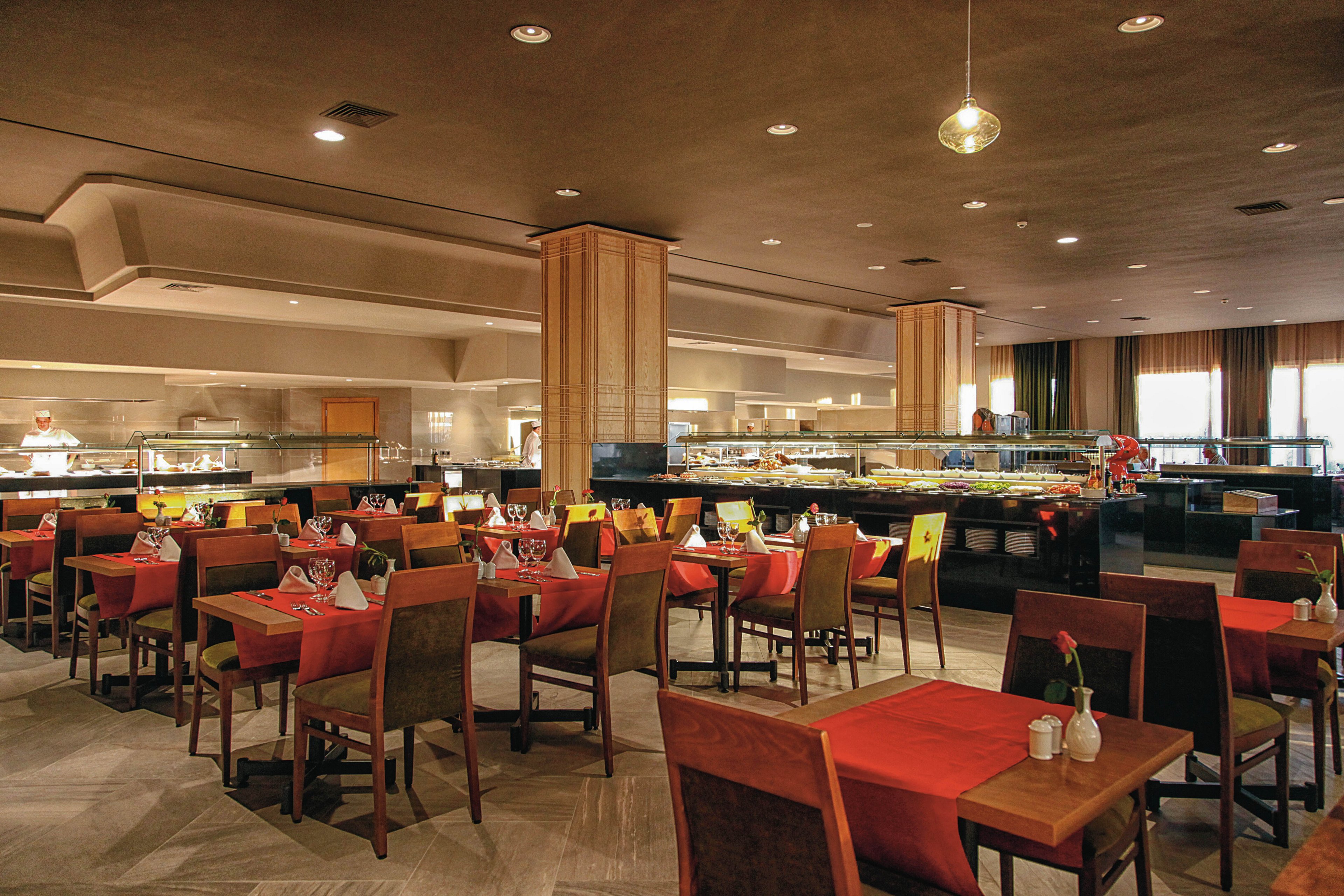 The image shows a large, modern buffet restaurant with neatly arranged tables covered in red tablecloths. The setting features warm lighting, wooden accents, and a variety of food stations in the background. Glassware and small vases with flowers are placed on each table, creating a welcoming and elegant atmosphere. The restaurant appears empty, highlighting its spacious layout and clean design.
