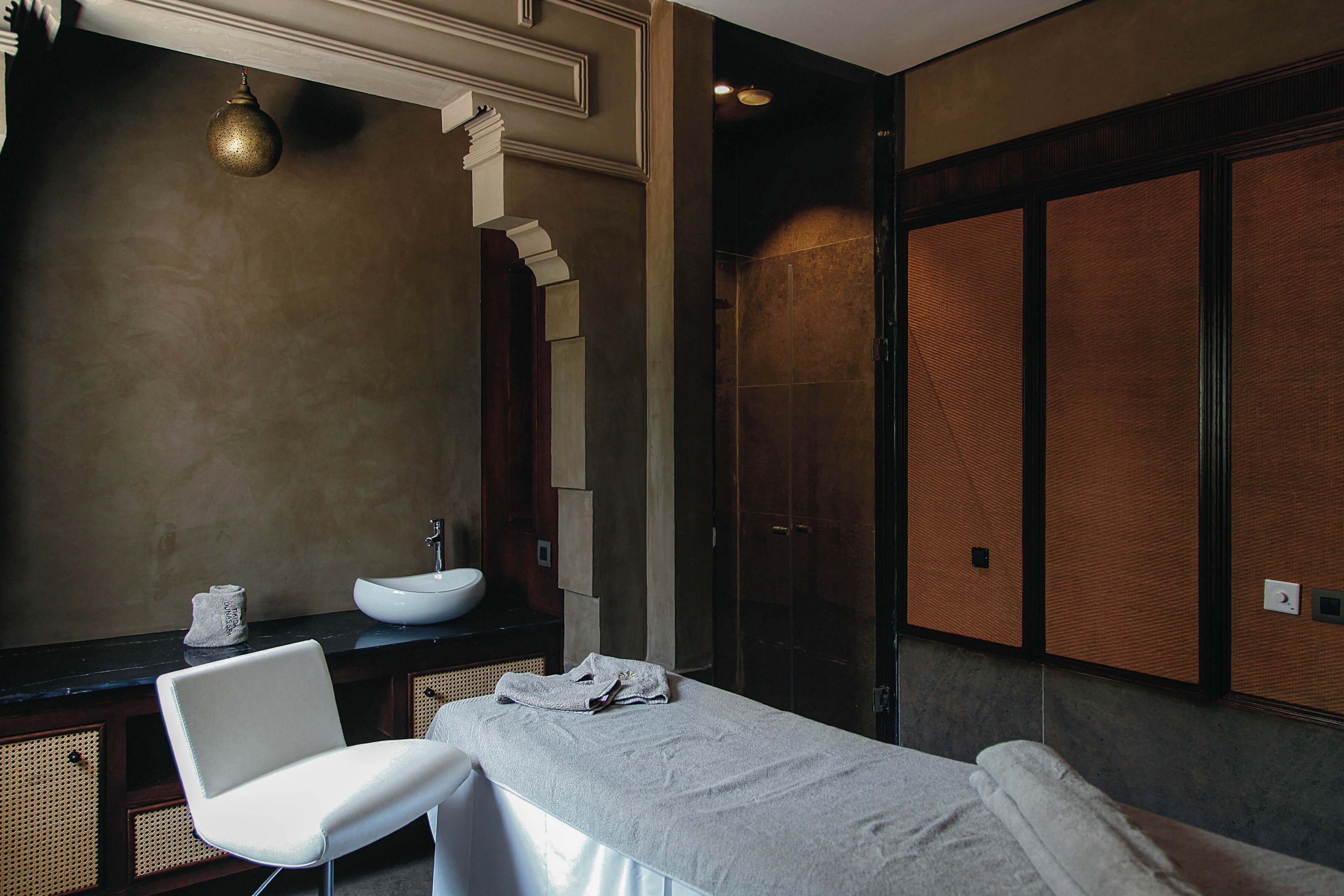 A contemporary spa treatment room features a massage table covered with a white sheet and towel. The setting includes a sleek white chair, a minimalist sink, and warm brown wall panels. The lighting is soft, creating a calm and relaxing atmosphere, with muted earth tones dominating the palette. No visible text or numeric values are present in the image.