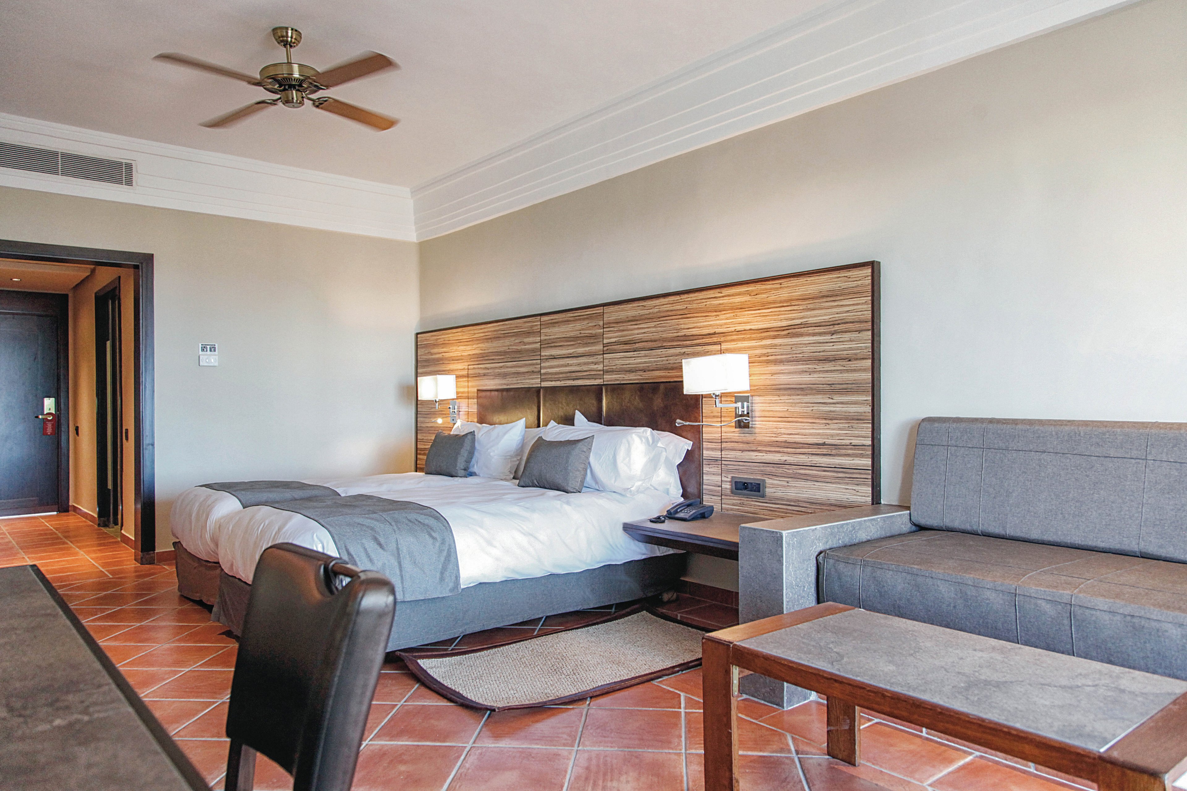 A spacious hotel room features a double bed with gray and white bedding, a wooden headboard, and two bedside lamps. The setting includes a gray sofa, wooden coffee table, and tiled floor, creating a clean and contemporary atmosphere. The room is well-lit, with neutral tones and minimalist decor, and a ceiling fan is visible above. No visible text or numbers are present in the image.