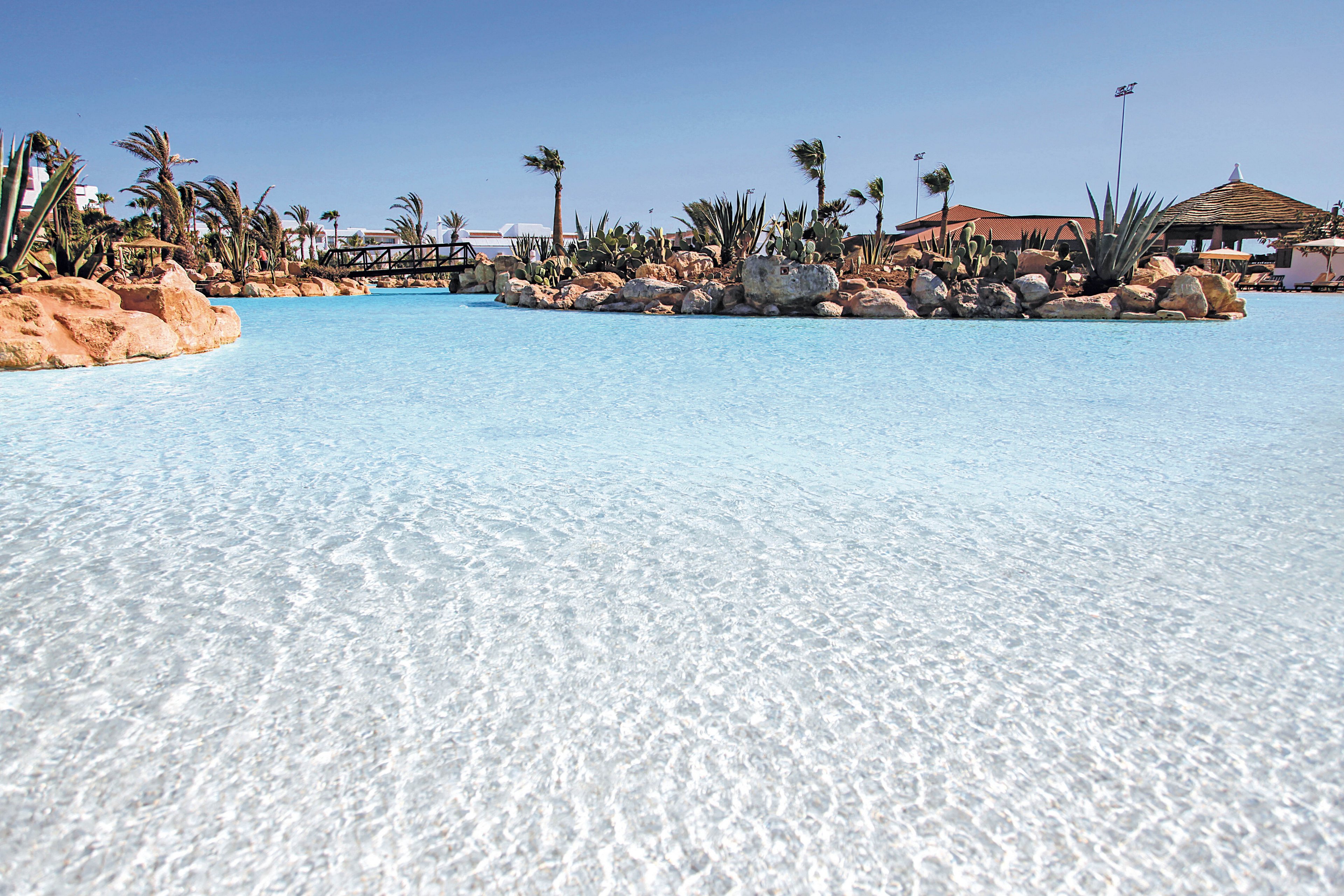 A large, clear lagoon pool is the main subject, surrounded by palm trees and rocky landscaping. The setting is an outdoor resort area under a bright blue sky, evoking a tropical and relaxing mood. The water is shallow and sparkling, with sandy textures visible beneath the surface. No people or visible text are present in the image.