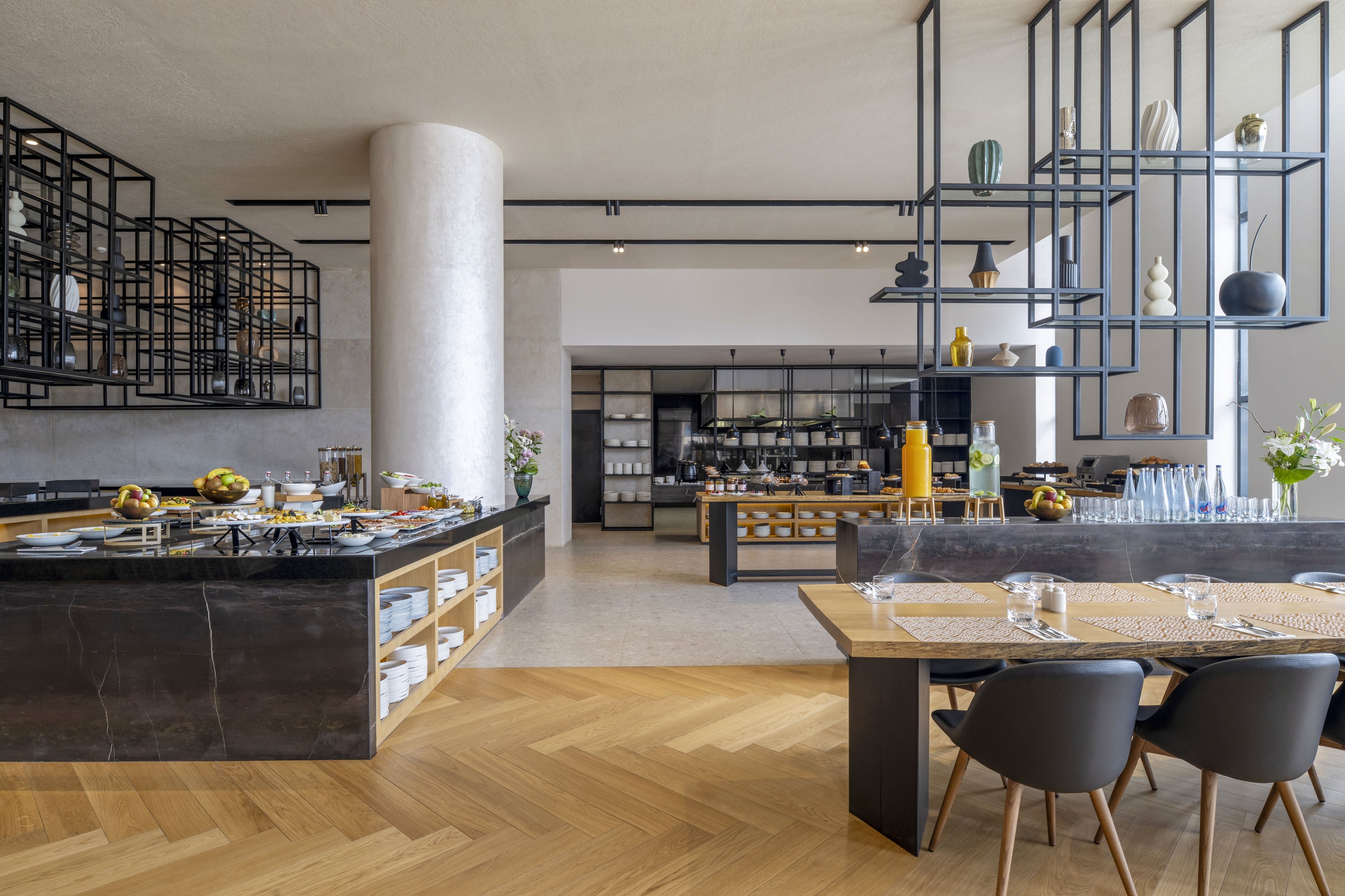 Contemporary buffet restaurant interior featuring a long marble counter with assorted dishes and serving platters. A spacious open kitchen is visible in the background with shelves, cabinets, and cooking equipment. Wooden herringbone flooring and light-colored walls create a warm, inviting atmosphere. Minimalist metal shelving with decorative vases and a neatly set dining table with chairs add to the modern design aesthetic.
