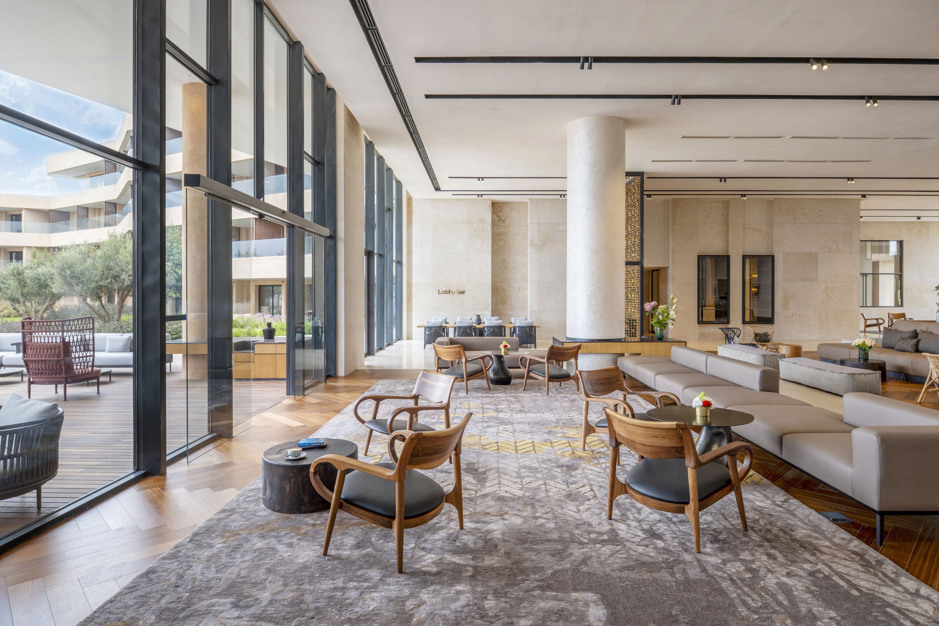 Spacious contemporary lobby interior features low sofas and wooden armchairs arranged for relaxed conversation. Floor-to-ceiling windows open to a terrace and landscaped courtyard, filling the space with natural light. Neutral tones, stone walls, and patterned carpet create a calm, upscale atmosphere. The setting suggests a modern hotel or resort lounge designed for comfort and socializing.