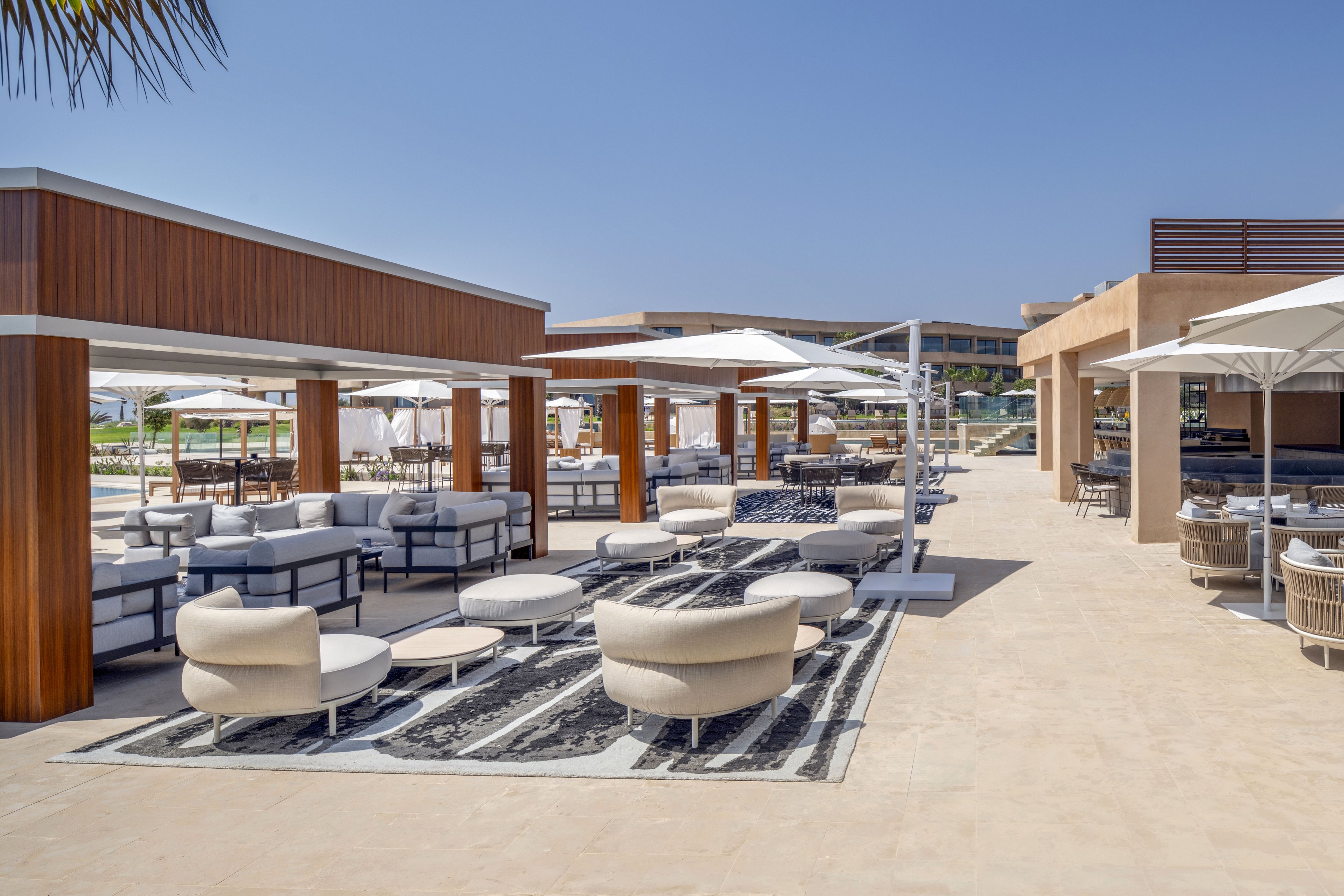 A contemporary outdoor lounge area features cushioned seating and round ottomans arranged beneath large white umbrellas. Wooden cabana-style structures frame the space, creating shaded zones for relaxation on a patterned rug. The setting appears to be part of a resort or hotel complex, with additional buildings and greenery visible in the background. Bright daylight and clear blue sky contribute to a clean, luxurious and inviting atmosphere.