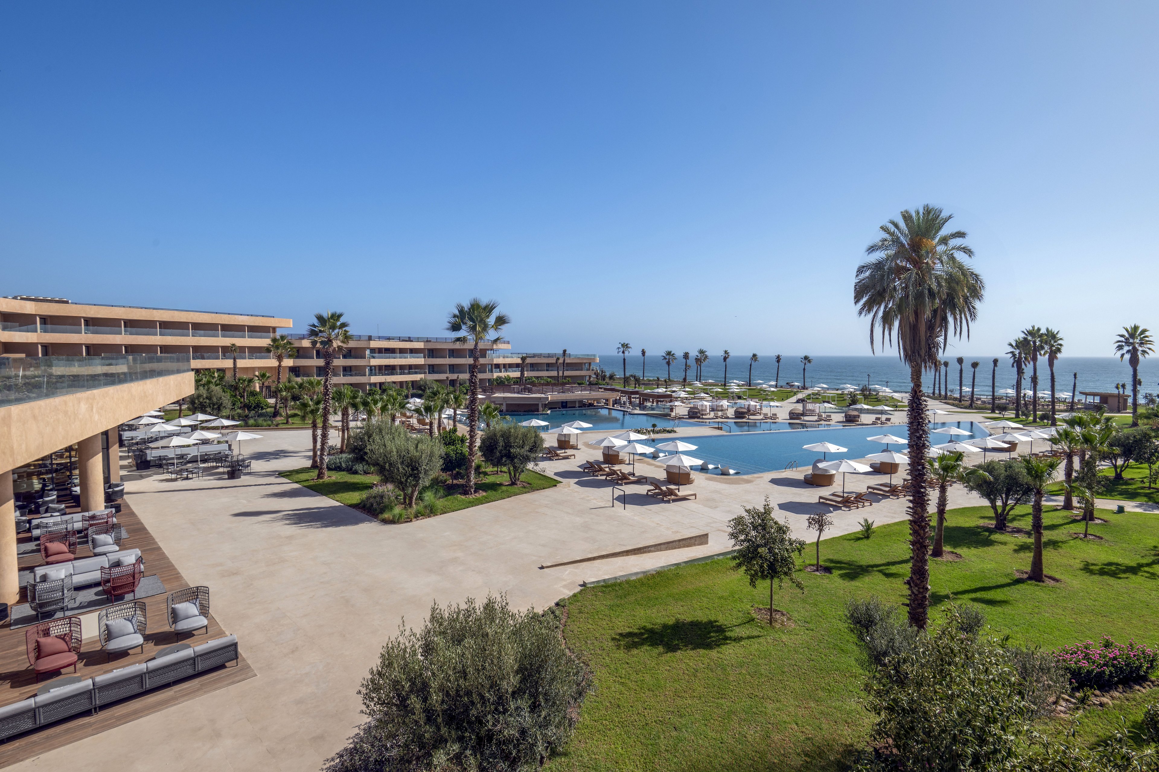 A large coastal resort features an expansive outdoor pool surrounded by sun loungers and white umbrellas. The scene includes manicured green lawns, palm trees, and landscaped gardens leading toward the shoreline and ocean horizon. Modern low-rise buildings frame the pool area, with shaded seating terraces overlooking the central courtyard. Clear blue sky and bright sunlight create a relaxed, vacation atmosphere at this beachfront property.