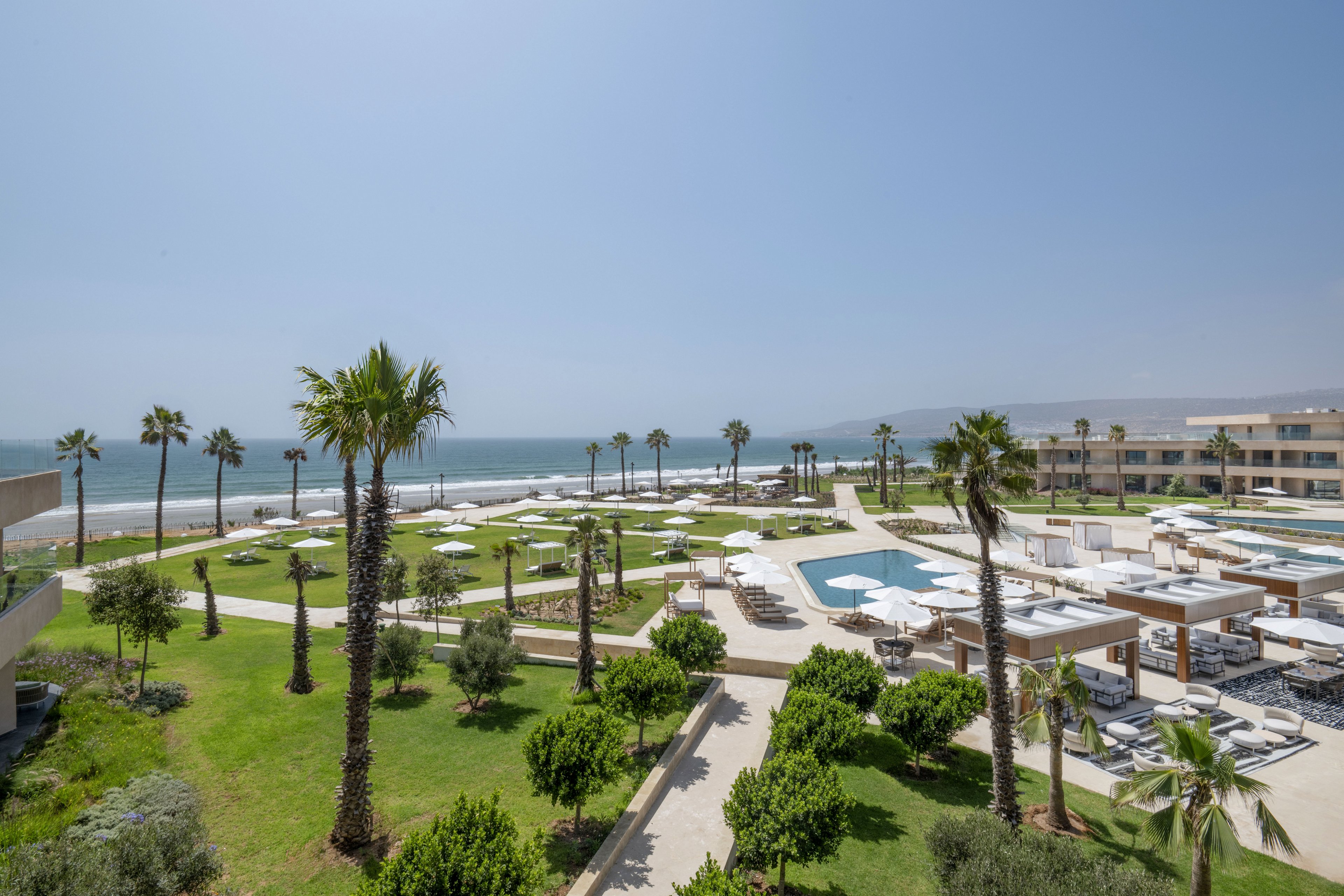 A modern beachfront resort overlooks the ocean with manicured lawns and tall palm trees. White sun loungers and umbrellas surround multiple outdoor pools and seating areas. Contemporary low-rise buildings frame the scene, creating a spacious, open layout. Clear blue sky and calm sea emphasize a relaxed, vacation atmosphere.
