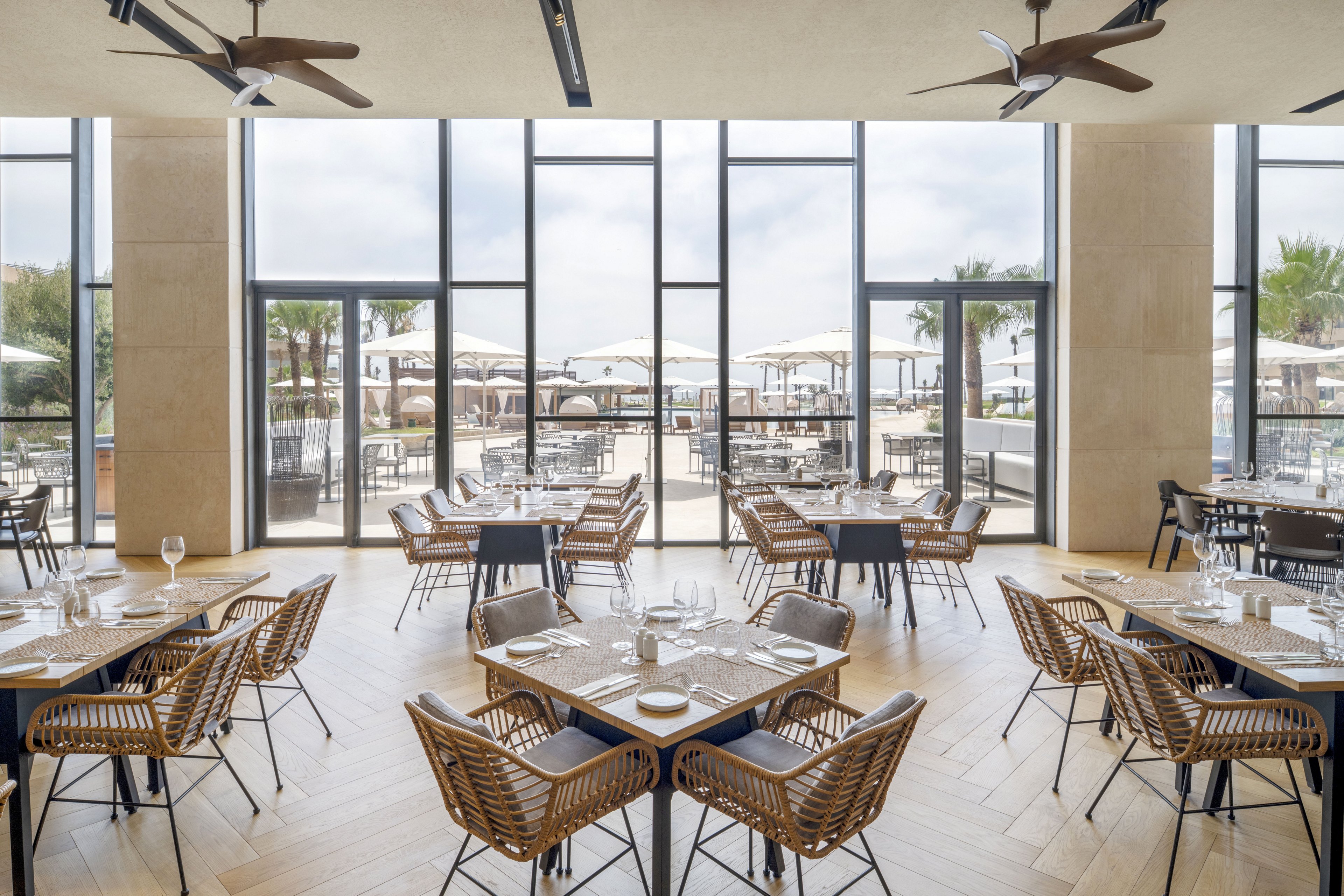 A contemporary restaurant dining room features neatly arranged tables and wicker chairs facing large floor-to-ceiling windows. The bright interior opens onto an outdoor terrace with umbrellas and additional seating, suggesting a resort or beachfront setting. Natural light floods the space, highlighting the light wood flooring and neutral-toned walls. The overall atmosphere feels airy, relaxed, and inviting for guests.