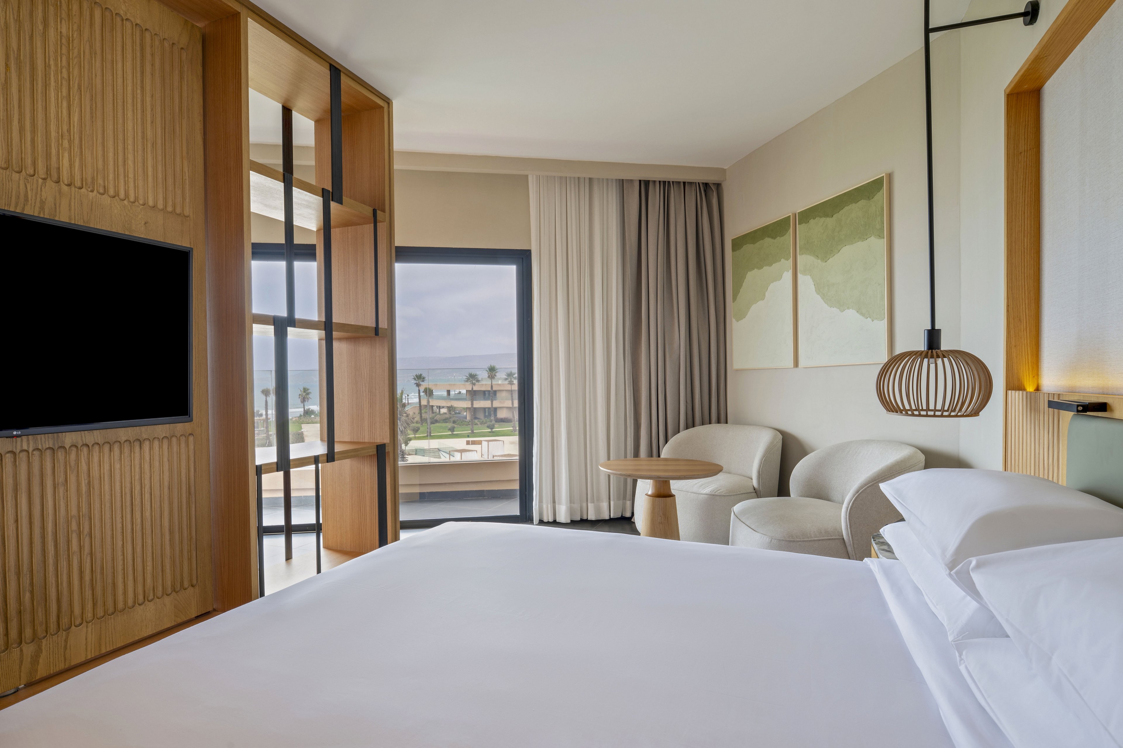 A contemporary hotel room features a neatly made bed facing a wall-mounted television and a balcony. Large sliding glass doors and sheer curtains open to an outdoor view of palm trees and coastal buildings. Soft, neutral-toned armchairs, a small round table, and abstract landscape artwork create a calm, minimalist atmosphere. Warm wood accents and a sculptural pendant light enhance the relaxed, resort-like setting.