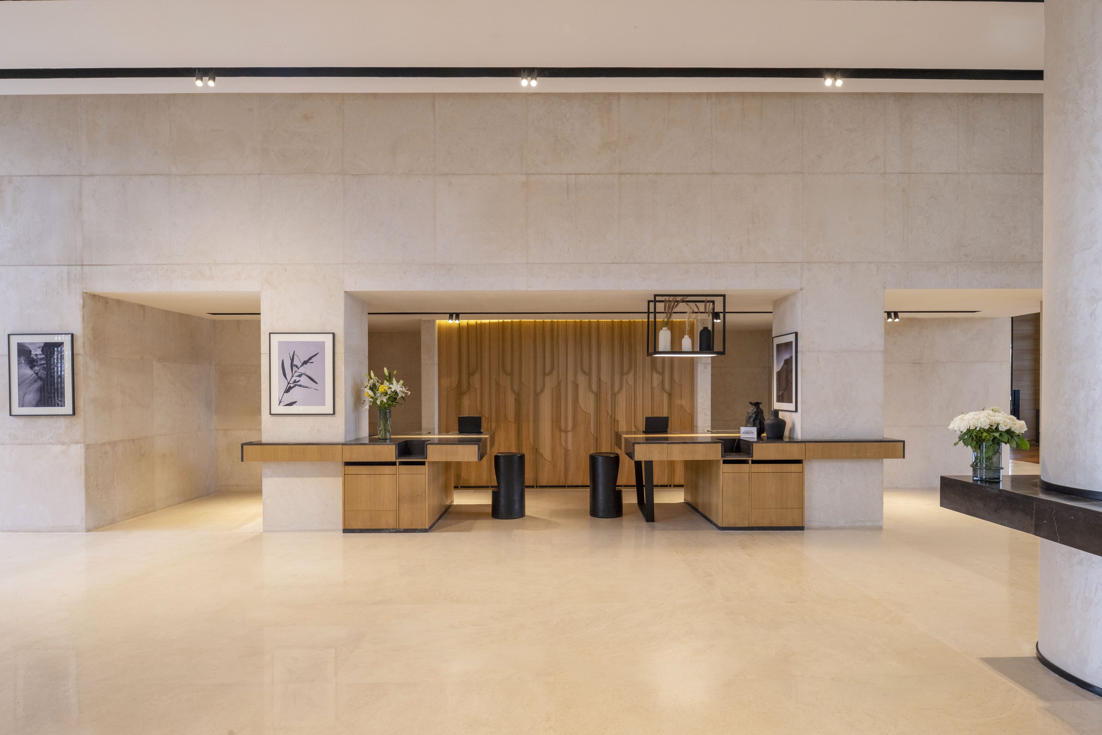A spacious, modern hotel reception lobby features a central wooden front desk with clean, minimalist lines. Light stone walls and polished floors create an airy, neutral environment. Framed artwork, fresh flower arrangements, and warm recessed lighting add subtle decorative accents. The overall scene is calm and uncluttered, emphasizing contemporary design and hospitality.