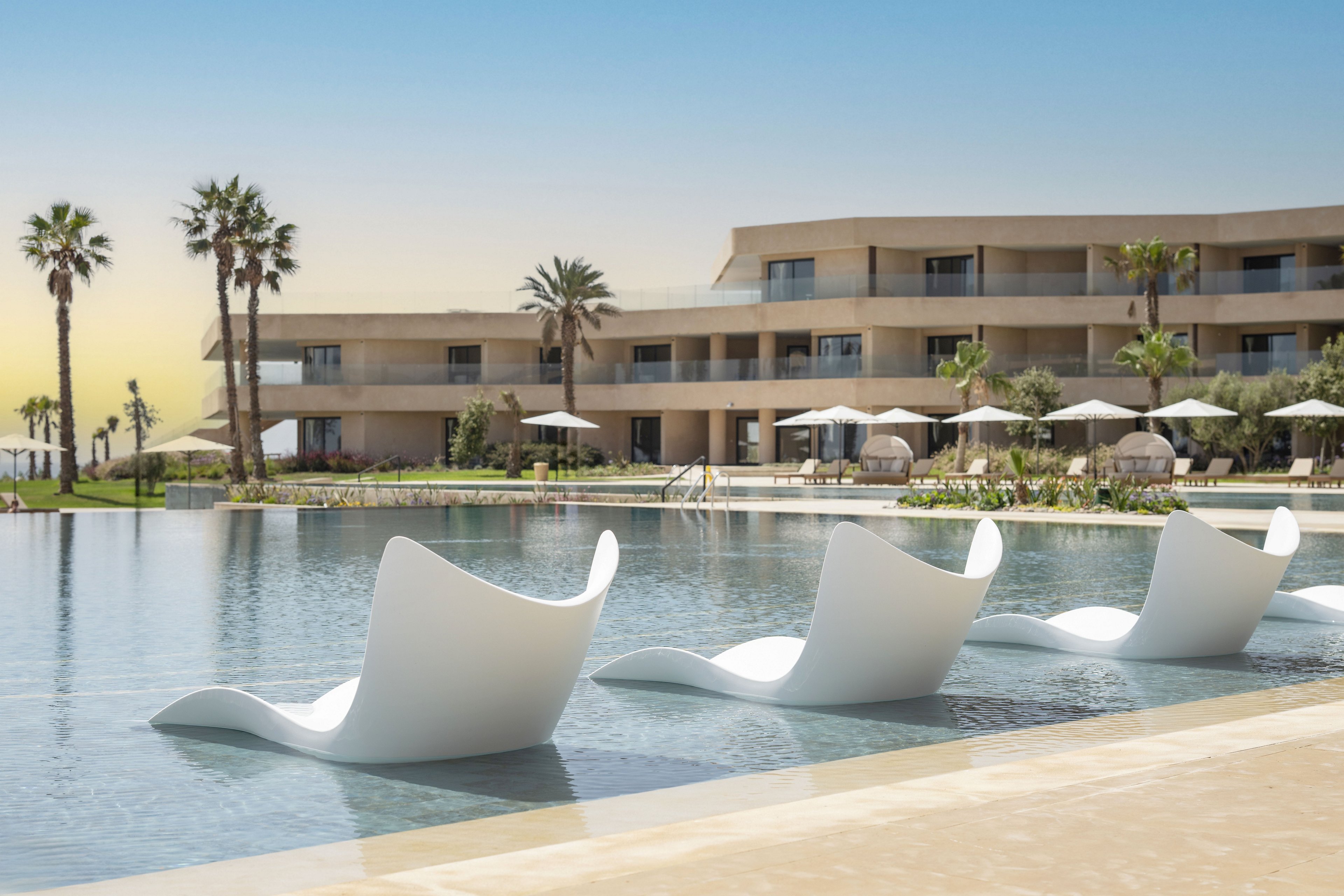 A contemporary resort features a tranquil infinity-style pool with sculptural white loungers partially submerged in the shallow water. In the background, a multi-story beige hotel building with balconies overlooks the pool area. Palm trees and neatly arranged sun umbrellas with lounge chairs line the pool deck, enhancing the vacation atmosphere. The sky is clear with a warm gradient, suggesting a sunny, relaxing day at a luxury destination.