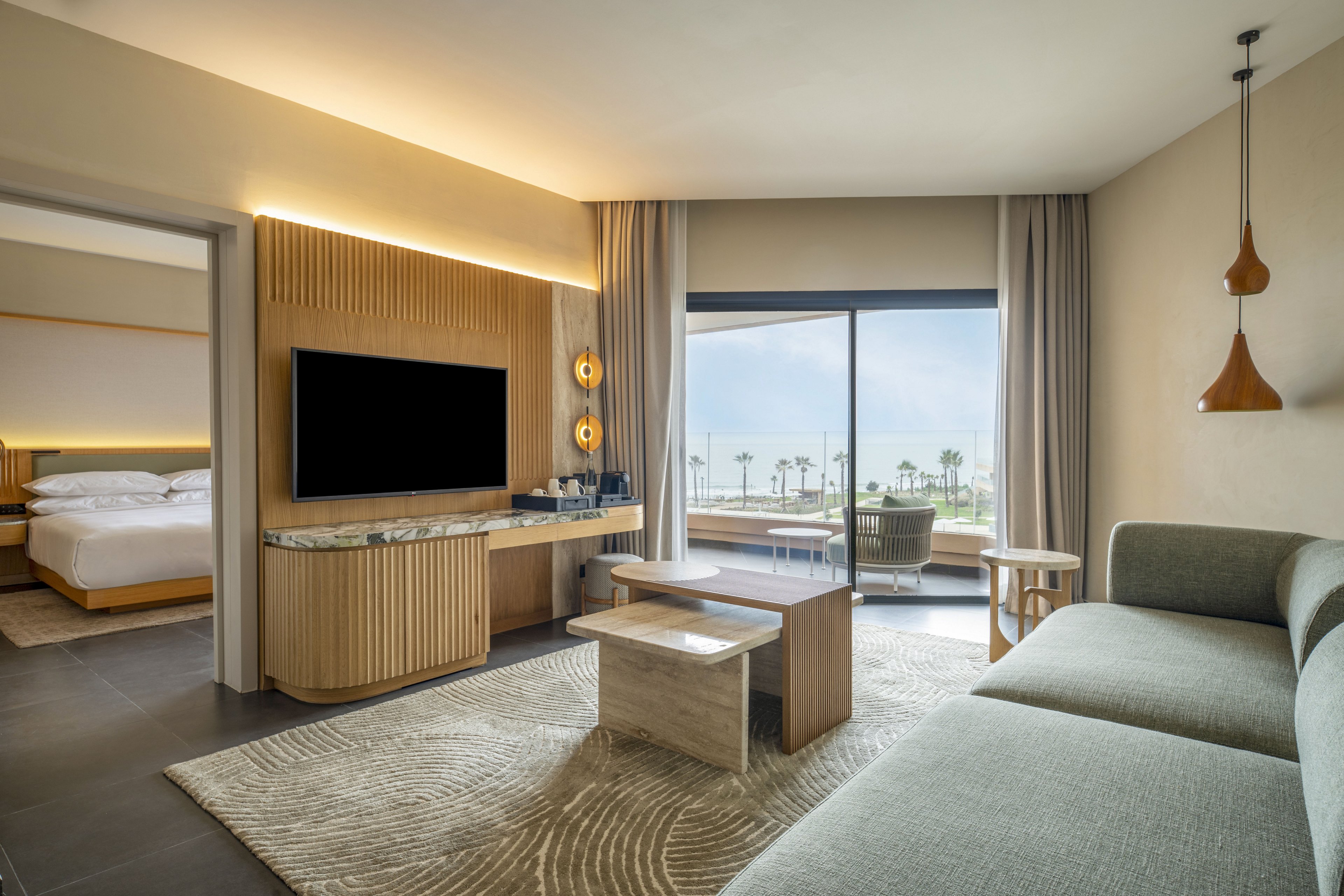 Contemporary hotel suite featuring a spacious living area with a sofa, nested coffee tables, and a large wall-mounted television. Warm wood accents and soft neutral tones create a calm, upscale atmosphere. A sliding glass door opens to a balcony with seating and views of palm trees and the ocean. An adjoining bedroom with a neatly made bed is visible through an open doorway, enhancing the sense of space and comfort.