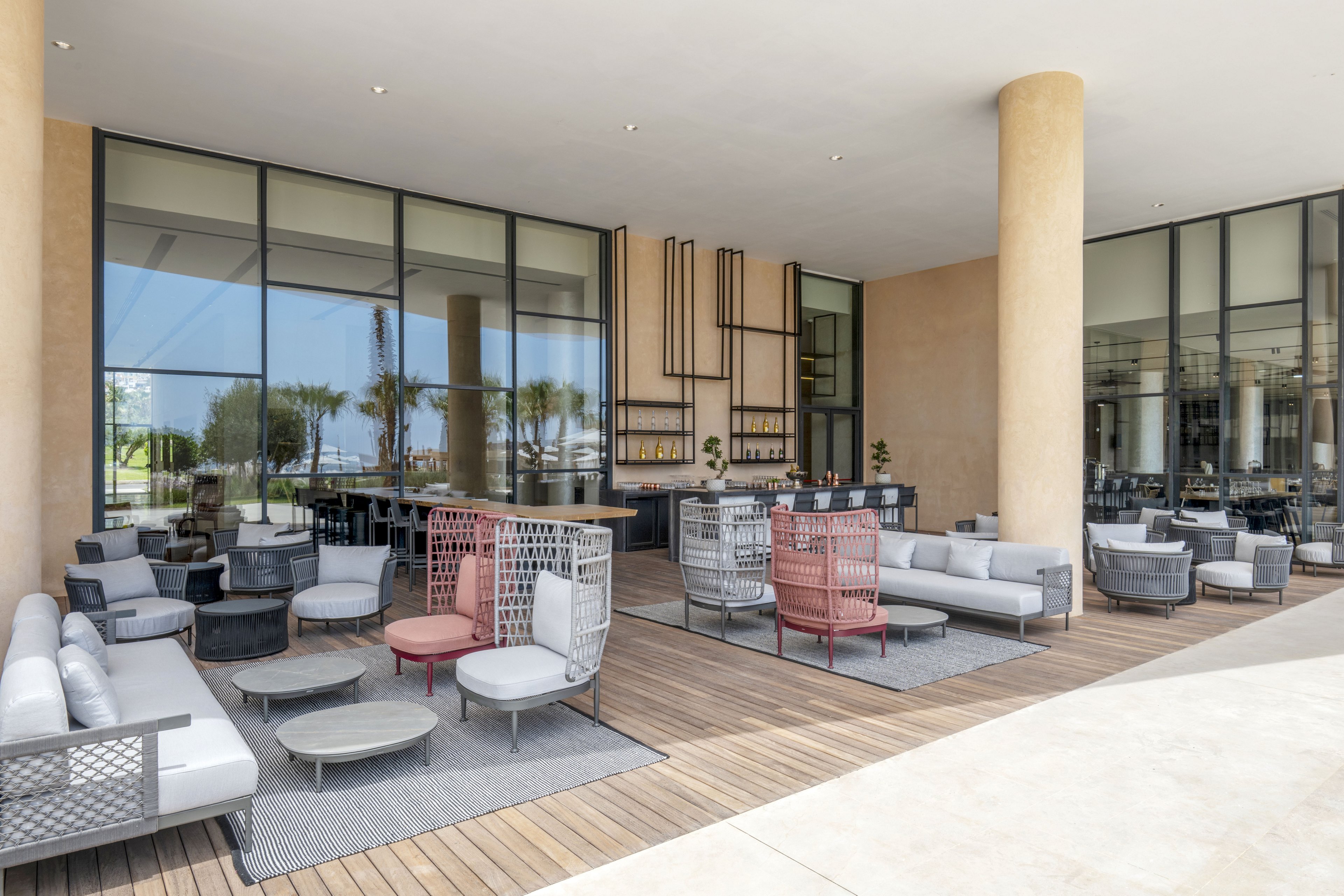 A contemporary outdoor lounge area features cushioned sofas and armchairs arranged on a wooden deck. Large floor-to-ceiling windows and tall beige columns frame the open-air space. Neutral gray tones are accented by muted red chairs and minimalist wall decor behind a sleek bar counter. The setting appears calm and inviting, ideal for relaxing or socializing in a resort or hotel environment.