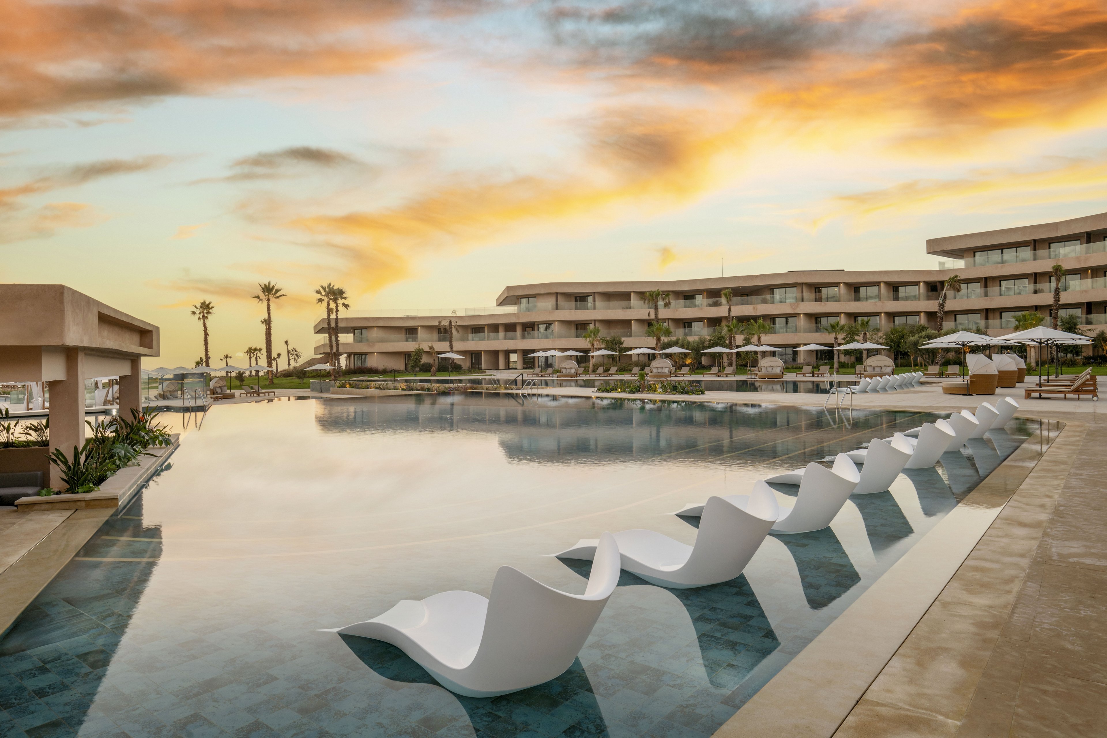 A contemporary resort features a large infinity-style swimming pool lined with white lounge chairs partially submerged in the water. Low-rise hotel buildings surround the pool, with balconies facing the central courtyard. Palm trees and neatly arranged sun umbrellas add to the vacation atmosphere. The sky is filled with warm sunset colors reflecting softly on the calm pool surface. The overall scene conveys relaxation and a luxurious holiday setting.