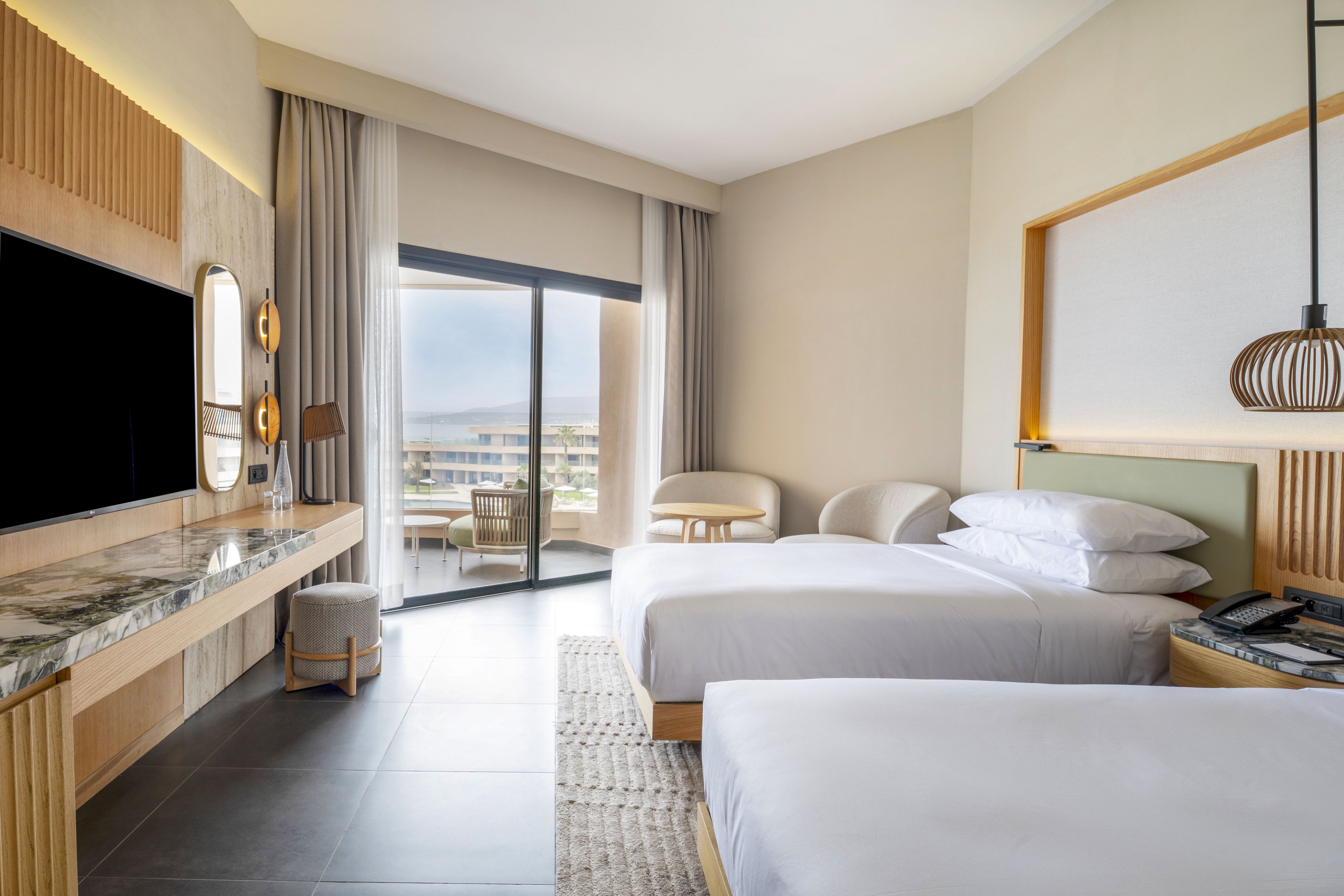 Contemporary hotel guest room featuring two neatly made twin beds with white linens. A large flat-screen TV is mounted above a marble-topped desk opposite the beds. Floor-to-ceiling sliding glass doors open to a balcony with outdoor seating and a view of nearby buildings and water. Neutral tones, soft lighting, and minimalist furniture create a calm, upscale atmosphere.