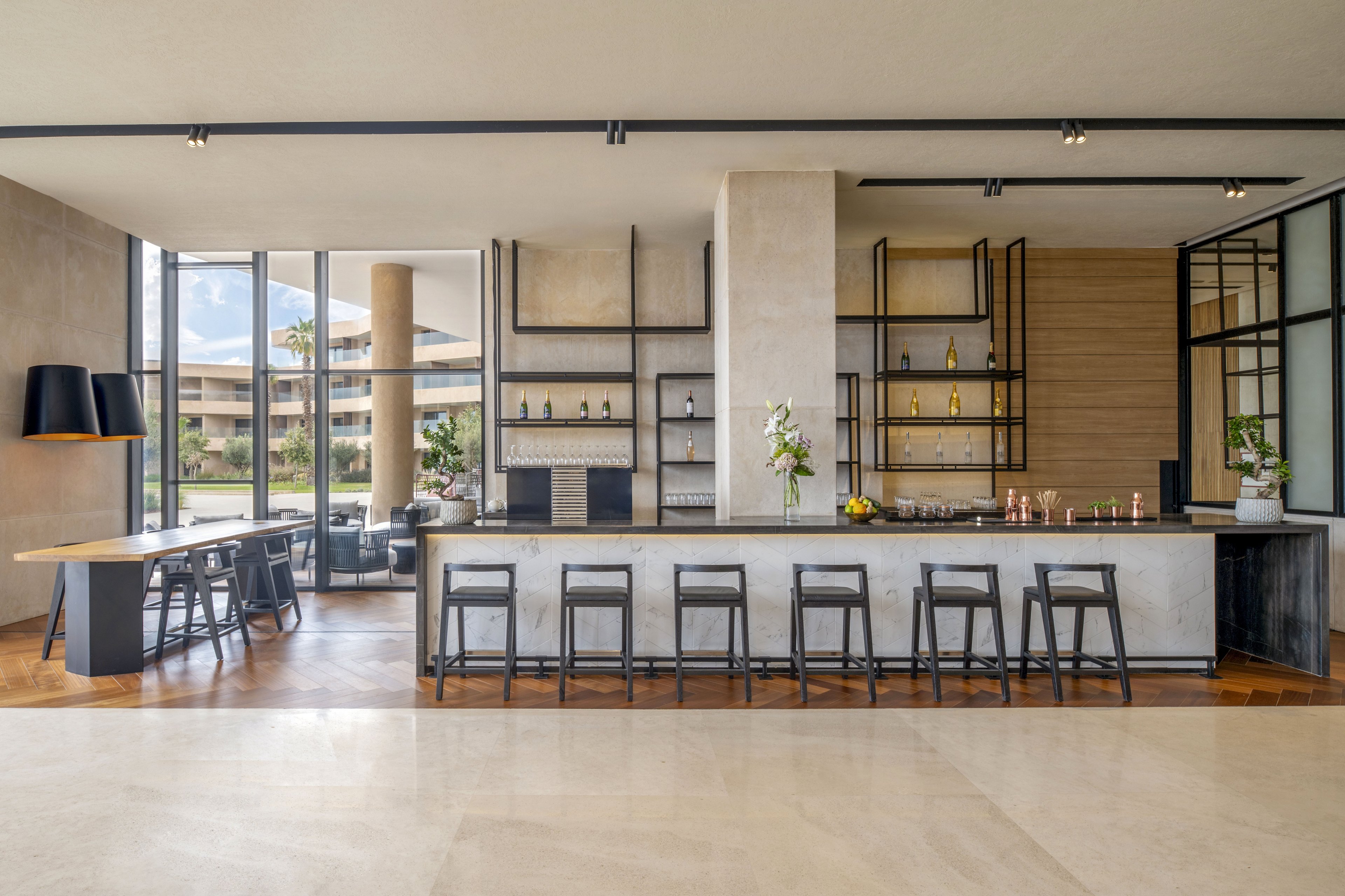 A contemporary hotel bar features a long marble counter with several barstools neatly arranged in front. Floor-to-ceiling windows reveal an outdoor view and fill the space with natural light. Minimalist shelving behind the bar displays bottles and decorative items, while a side table area offers additional seating. Neutral tones, clean lines, and polished surfaces create a sophisticated and inviting atmosphere.