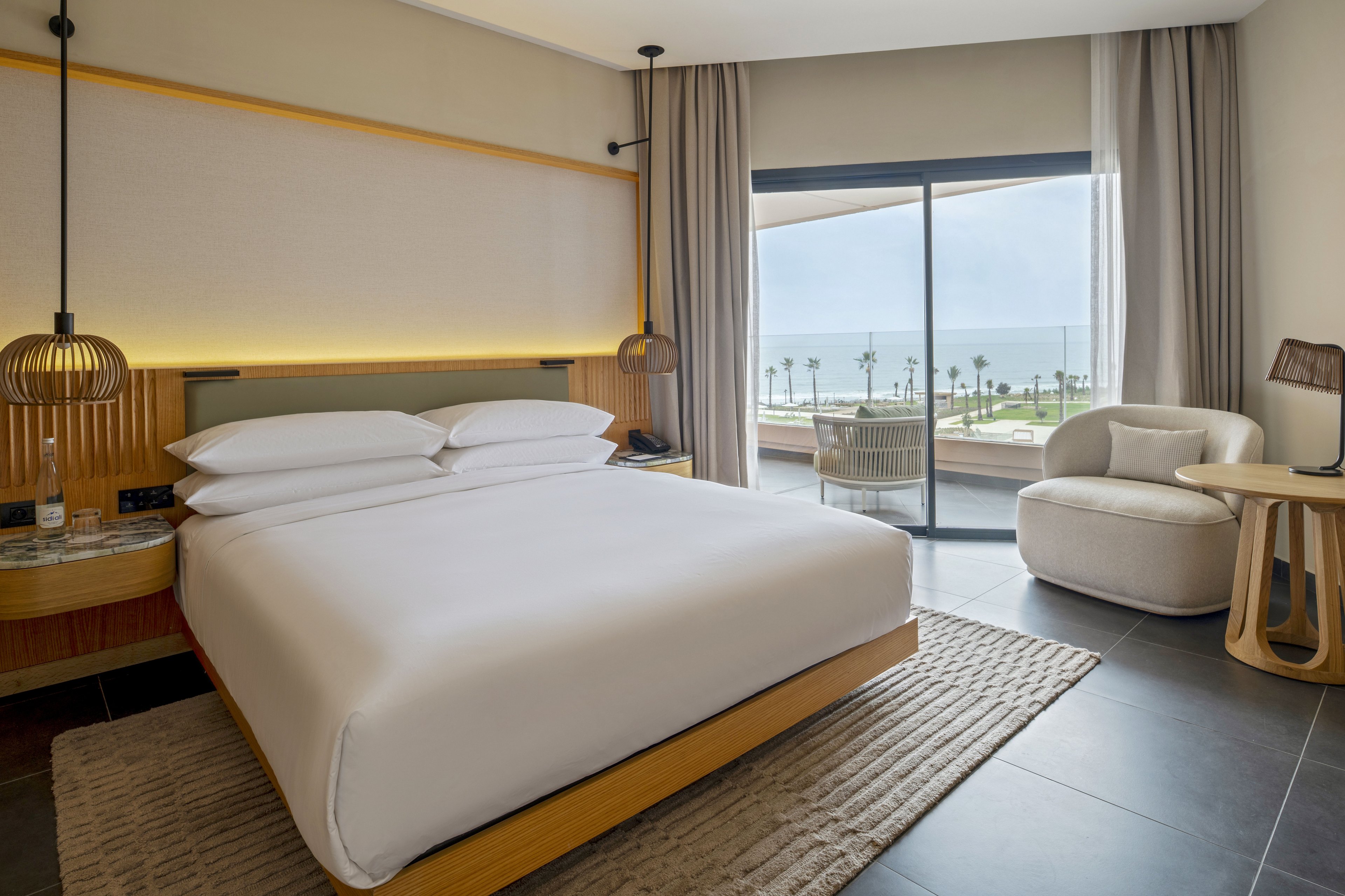 Contemporary hotel room featuring a large bed with crisp white linens and wooden frame. The space includes bedside pendant lamps, a textured rug, and a cozy armchair with a small side table. Floor-to-ceiling glass doors open to a balcony overlooking palm trees and the ocean. Neutral tones and soft lighting create a calm, minimalist atmosphere ideal for relaxation.