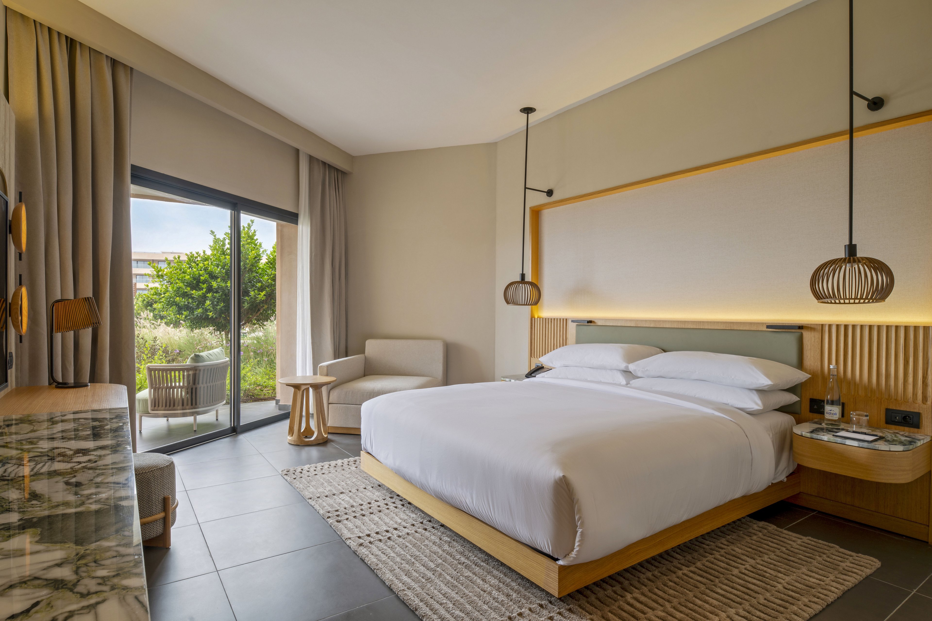 A contemporary hotel room features a large bed with crisp white linens and a wooden headboard. The space includes a lounge chair, small side tables, and pendant lights that create a warm, inviting atmosphere. A sliding glass door opens to an outdoor terrace with seating and greenery visible outside. Neutral tones, soft textures, and clean lines give the room a minimalist, upscale feel.