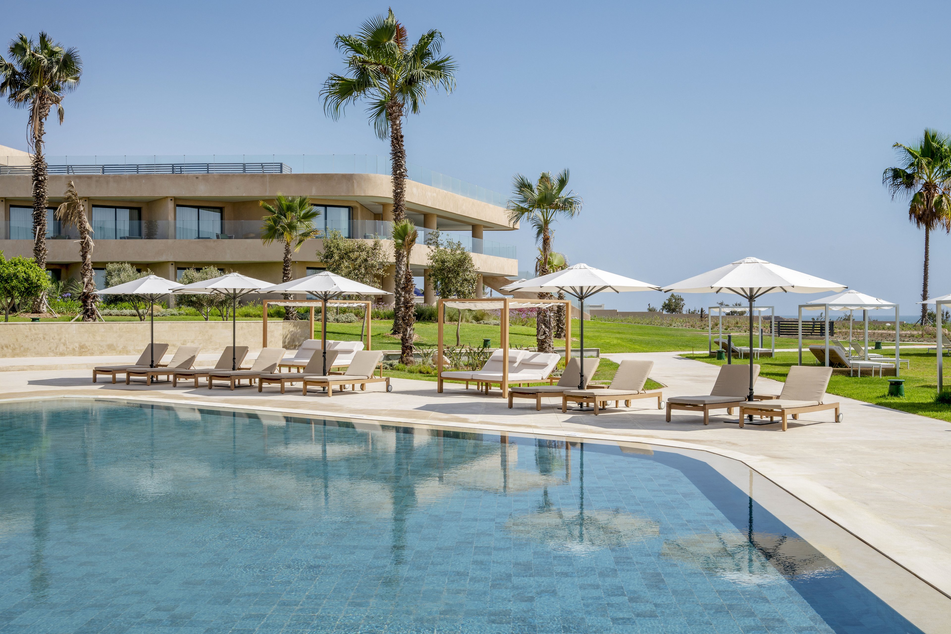 A serene outdoor swimming pool is surrounded by neatly arranged sun loungers and white parasols. In the background, a modern resort building with balconies overlooks a manicured lawn and palm trees. The clear blue sky and calm water create a relaxing vacation atmosphere. The scene suggests a high-end coastal resort designed for leisure and sunbathing.