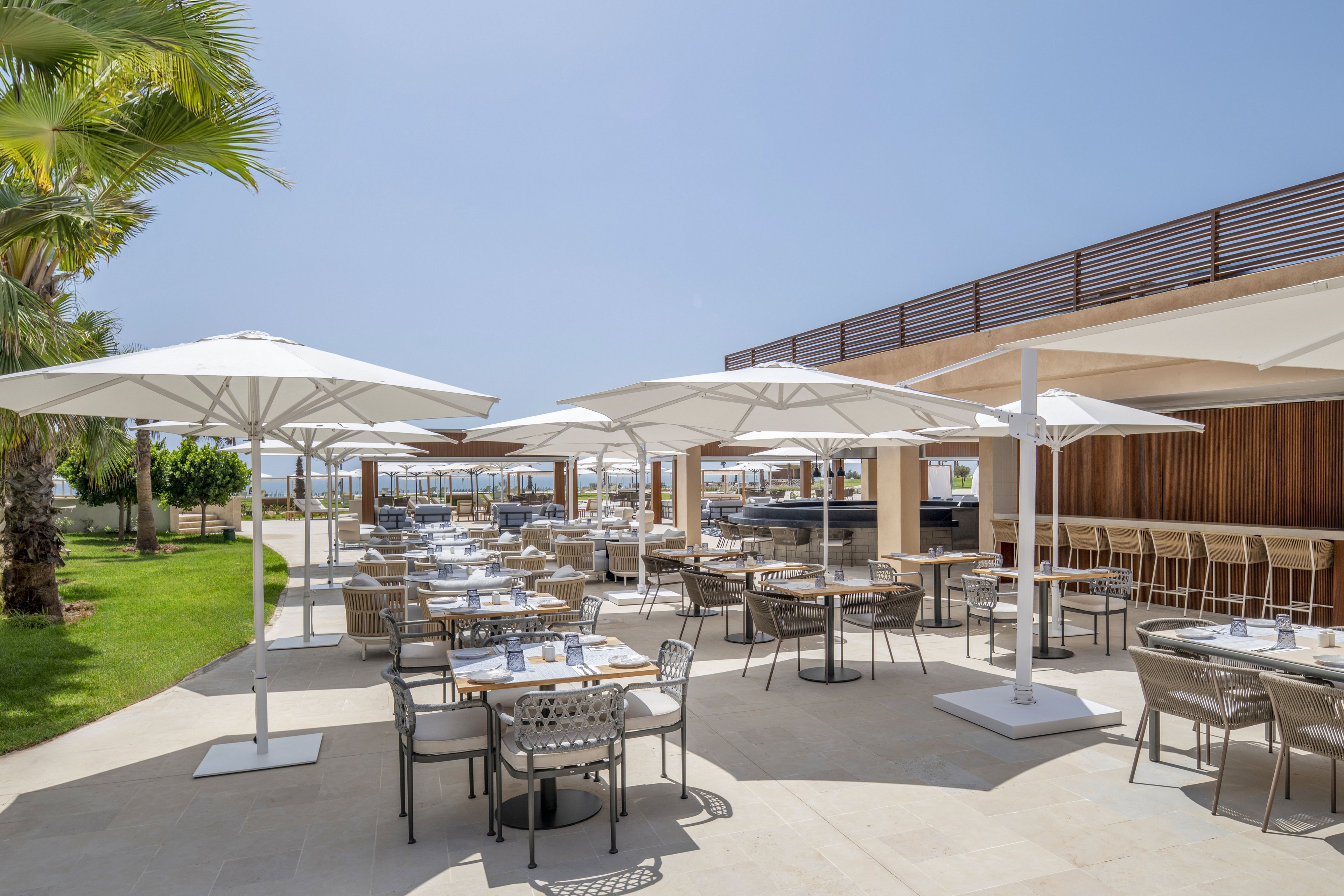 An open-air restaurant terrace is set with neatly arranged tables and chairs under large white umbrellas. The scene overlooks a bright seaside landscape with clear blue sky and palm trees. A long bar counter with high stools lines the right side, adding to the resort-like atmosphere. The overall setting feels relaxed and inviting, ideal for al fresco dining in warm weather.
