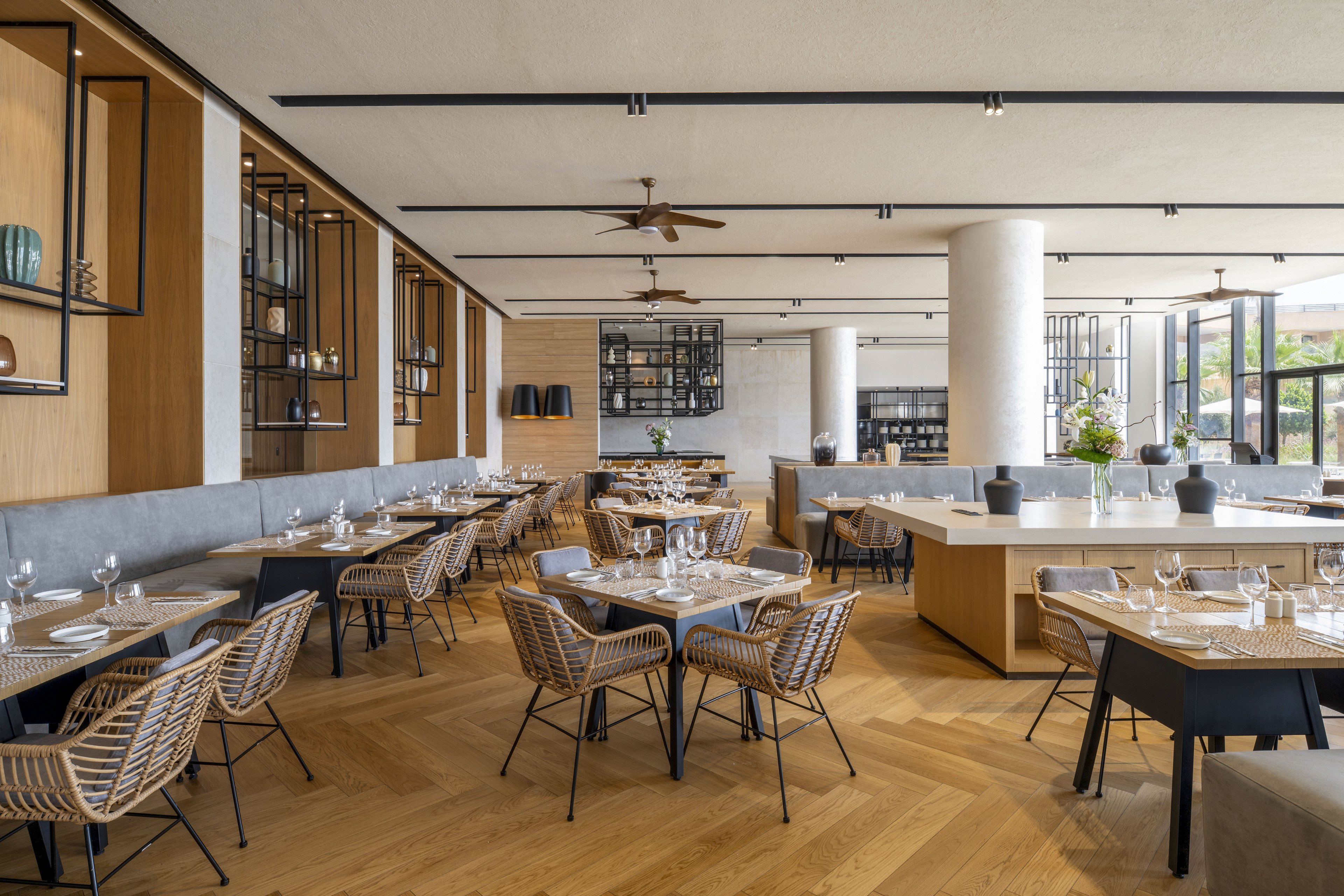 Contemporary restaurant dining room featuring wicker chairs and neatly set tables with glassware and plates. The space is open and bright, with large windows and light wood flooring creating an airy atmosphere. Minimalist shelving, ceiling fans, and neutral-toned benches contribute to a clean, modern design. The overall setting suggests a stylish, upscale yet relaxed dining experience.