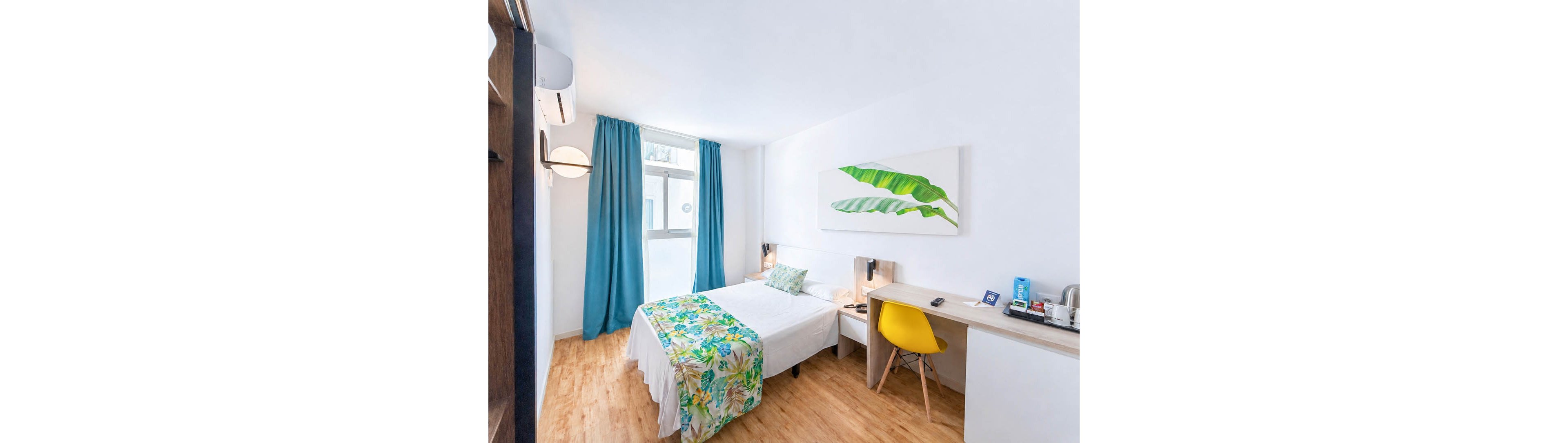 A bright, minimalist bedroom features a neatly made double bed with a tropical-patterned runner and pillow. The room has white walls, wood-look flooring, and a large window framed by teal curtains. A simple desk with a yellow chair holds a kettle, cups, and small amenities. A large leaf artwork above the bed adds a fresh, modern accent to the clean, airy space.