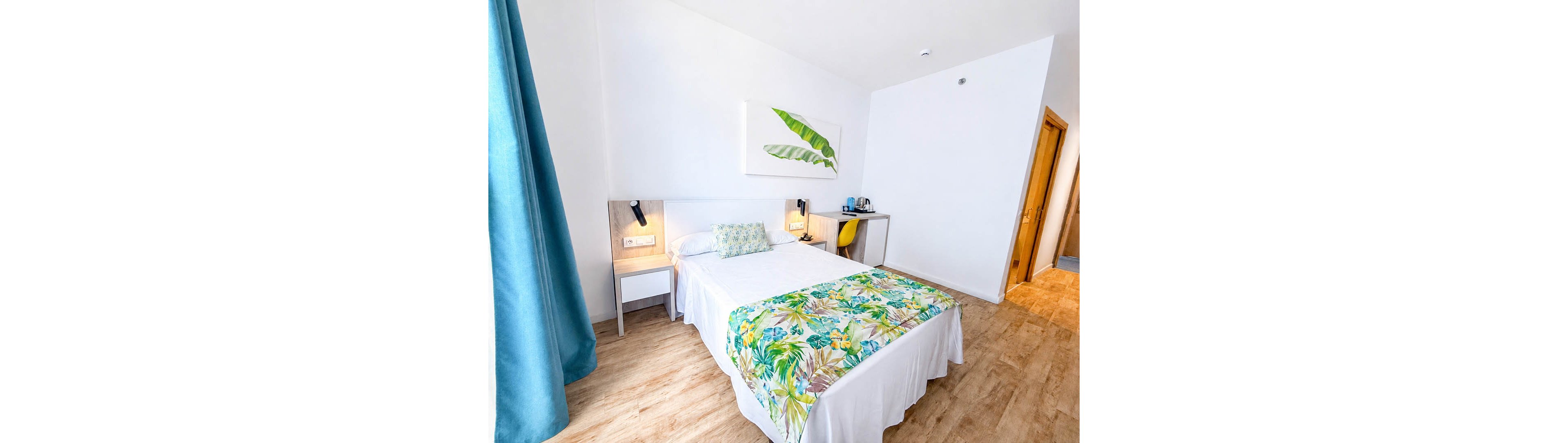 A neatly made double bed with white linens and a colorful tropical bed runner stands in a bright, modern room. The space features light wood flooring, white walls, and a large botanical artwork above the headboard. Bedside tables with wall-mounted reading lights flank the bed, while a desk with a yellow chair and a small beverage station sits to the side. Natural light enters through a window with blue curtains, creating an airy and inviting atmosphere.