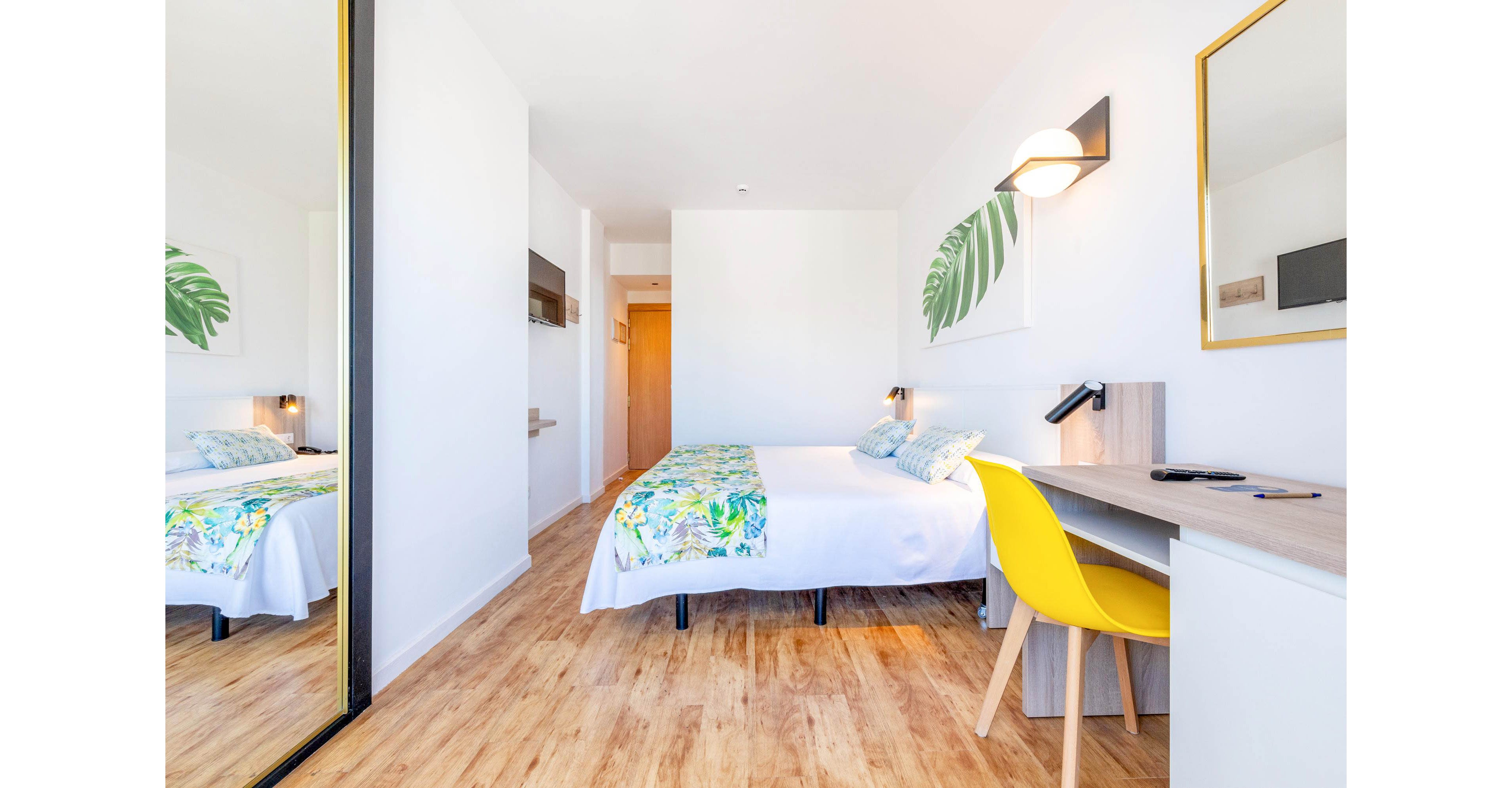 A bright minimalist bedroom features a neatly made bed with colorful tropical bedding. The room includes a wooden desk with a yellow chair and a wall-mounted lamp, creating a small workspace. A large mirror wardrobe reflects the space, enhancing the sense of openness. Light wooden flooring and white walls contribute to the airy, modern feel, complemented by simple leaf artwork above the bed.