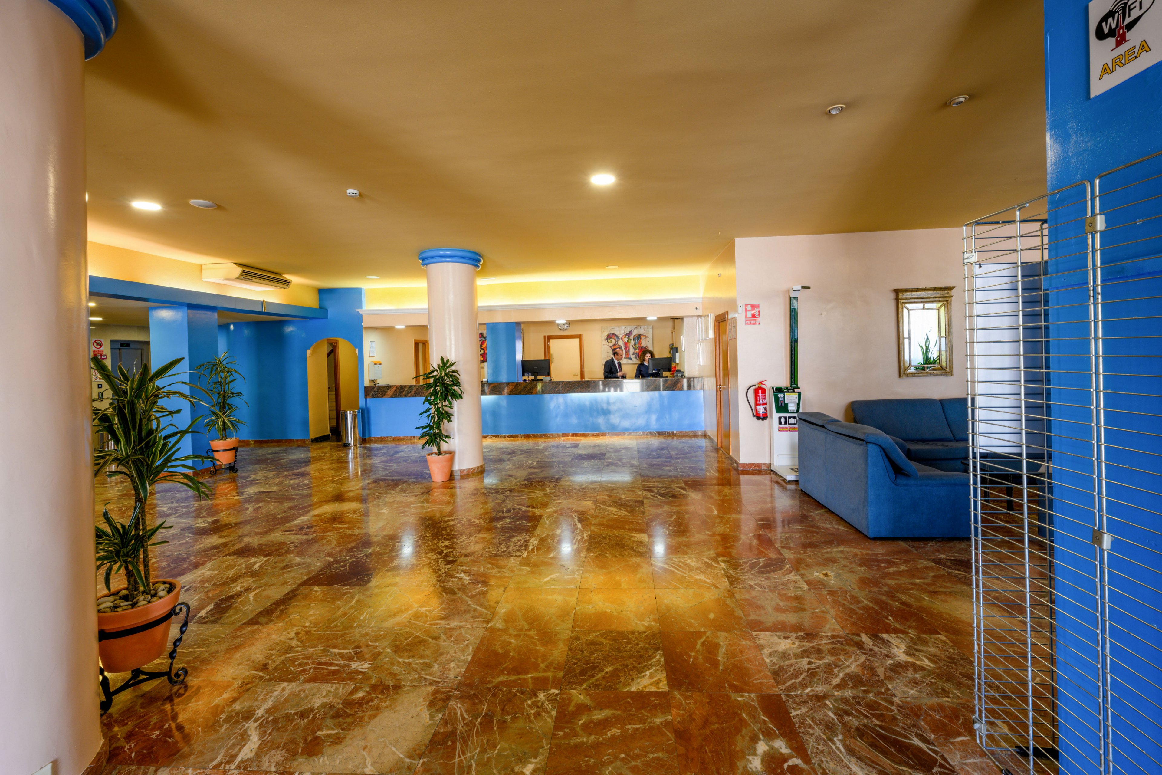 A wide hotel lobby features polished marble floors and pastel-colored walls. The reception desk with staff is visible in the background, framed by blue columns. Potted plants, a blue sofa seating area, and warm ceiling lighting create a welcoming atmosphere. The space appears clean, open, and ready to receive guests.