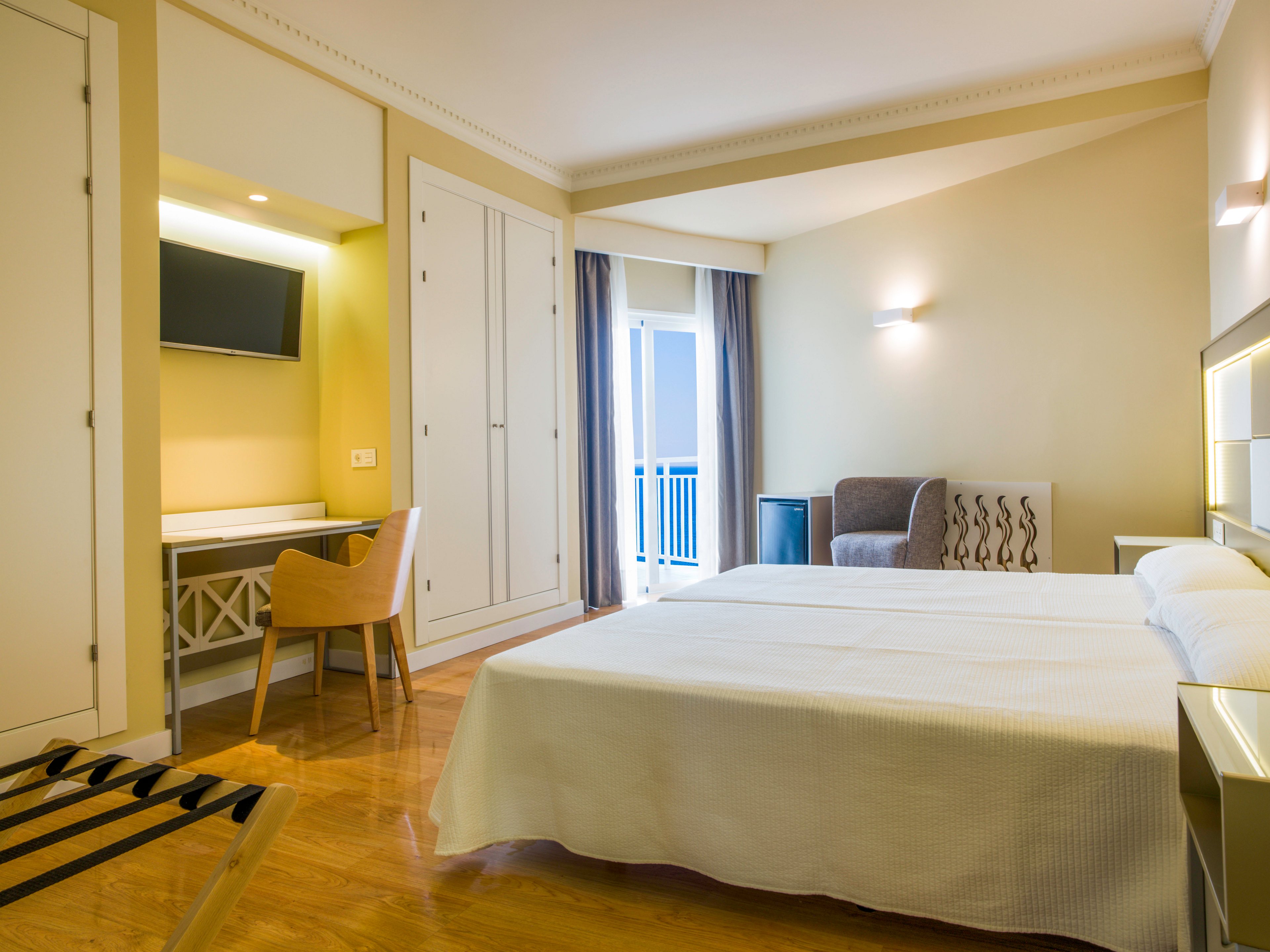 A spacious, modern hotel room features a neatly made double bed with white linens. The room includes built-in wardrobes, a wall-mounted flat-screen TV, and a small desk with a wooden chair. A cozy armchair sits near the balcony doors, which open to an outdoor view with blue railings. Warm lighting and light-colored walls create a calm, inviting atmosphere.