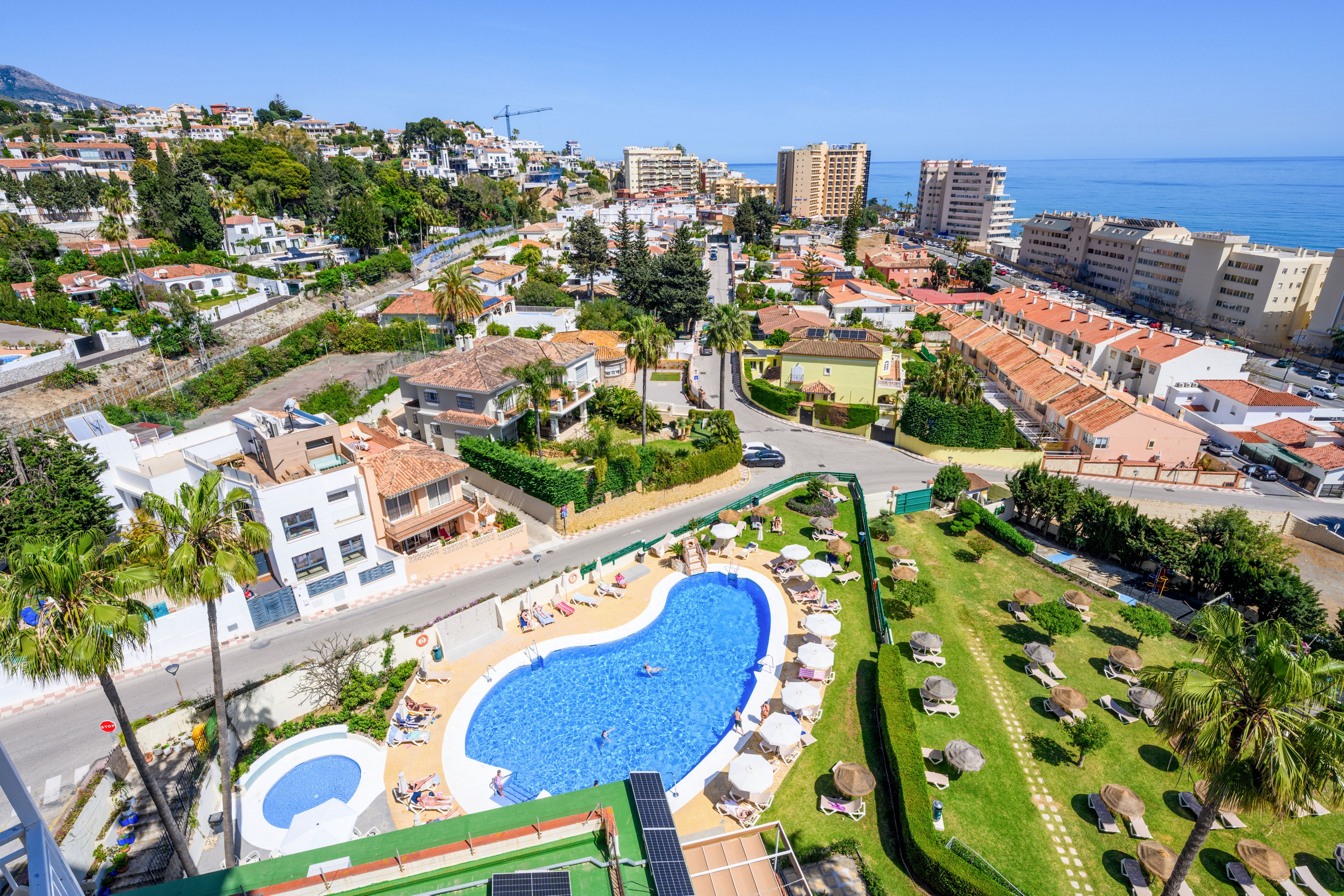 An aerial view shows a large outdoor swimming pool surrounded by sun loungers and parasols in a coastal resort. The pool area is bordered by manicured lawns, palm trees, and smaller circular pools. Residential buildings and hotels stretch toward the shoreline, with the sea visible in the background. The bright daylight and clear sky create a vibrant, summery atmosphere in this holiday destination.