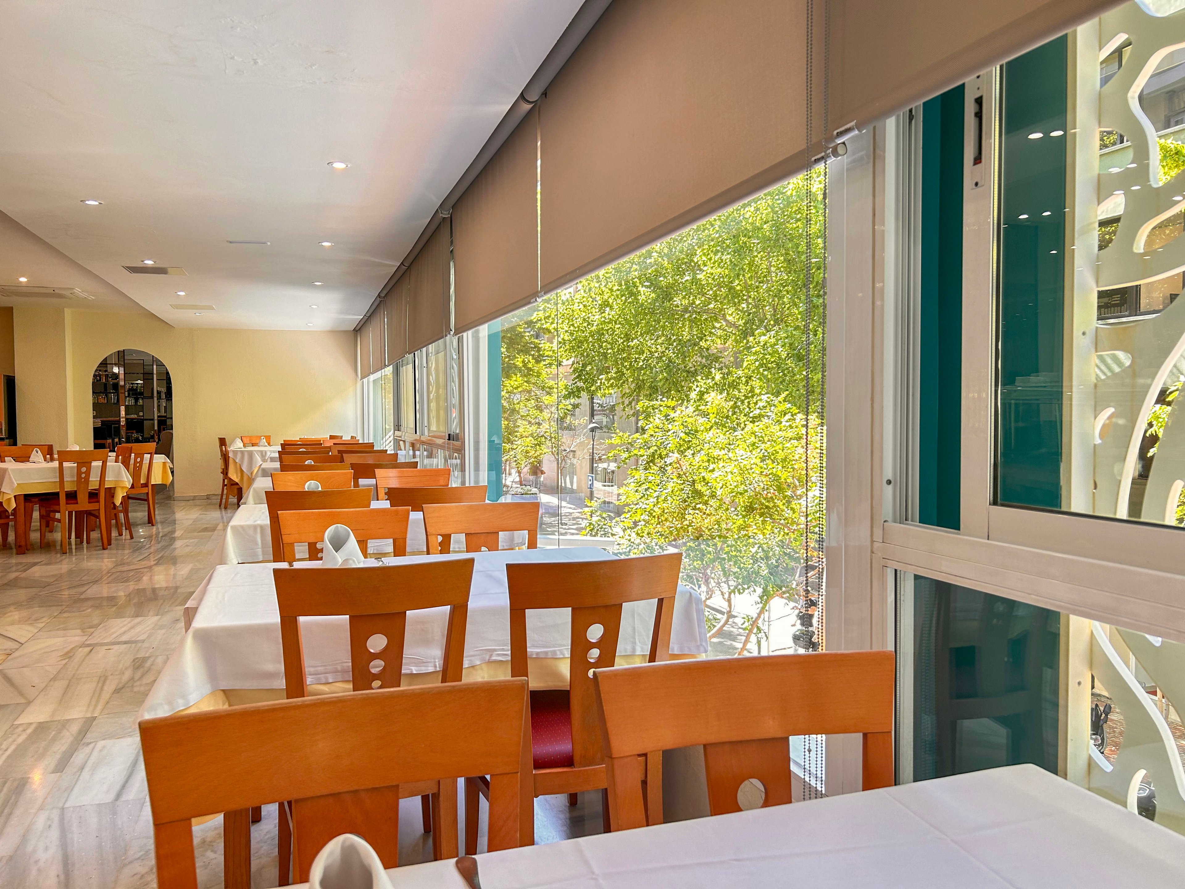 A sunlit restaurant dining area features neatly arranged wooden chairs and white tablecloths. Large floor-to-ceiling windows line one side, offering views of green trees and the street outside. The interior has a clean, modern look with light-colored walls and marble-style flooring. Tables are set but the space is currently empty, suggesting readiness for guests.