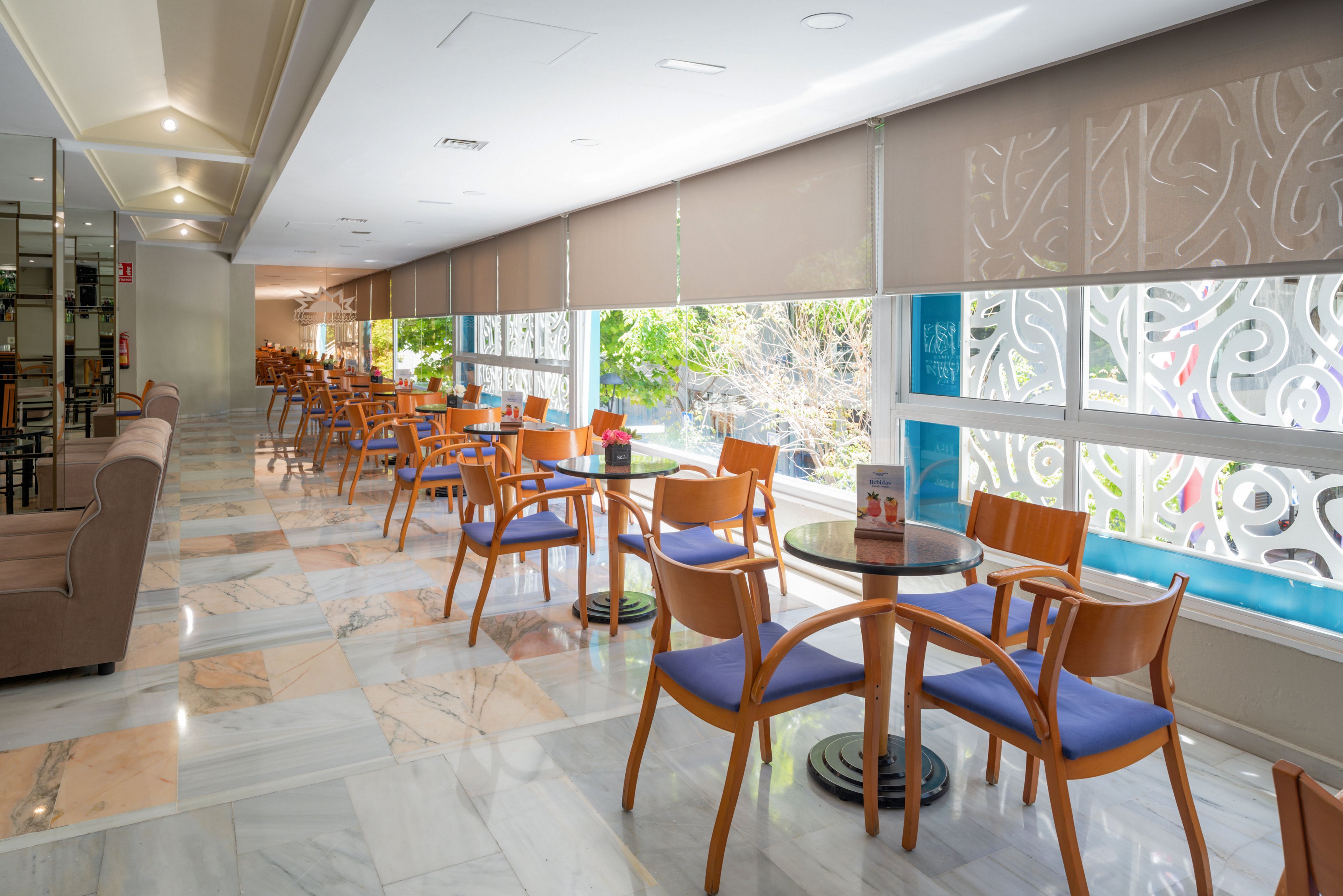 A sunlit cafe interior features rows of wooden chairs with blue cushions arranged around round glass-top tables. Large windows line one side, offering views of greenery outside and filtered light through decorative panels and roller blinds. The polished marble floor reflects the natural light, enhancing the clean and modern atmosphere. Menus and table accessories sit neatly on the tables, suggesting a ready-to-serve dining space.