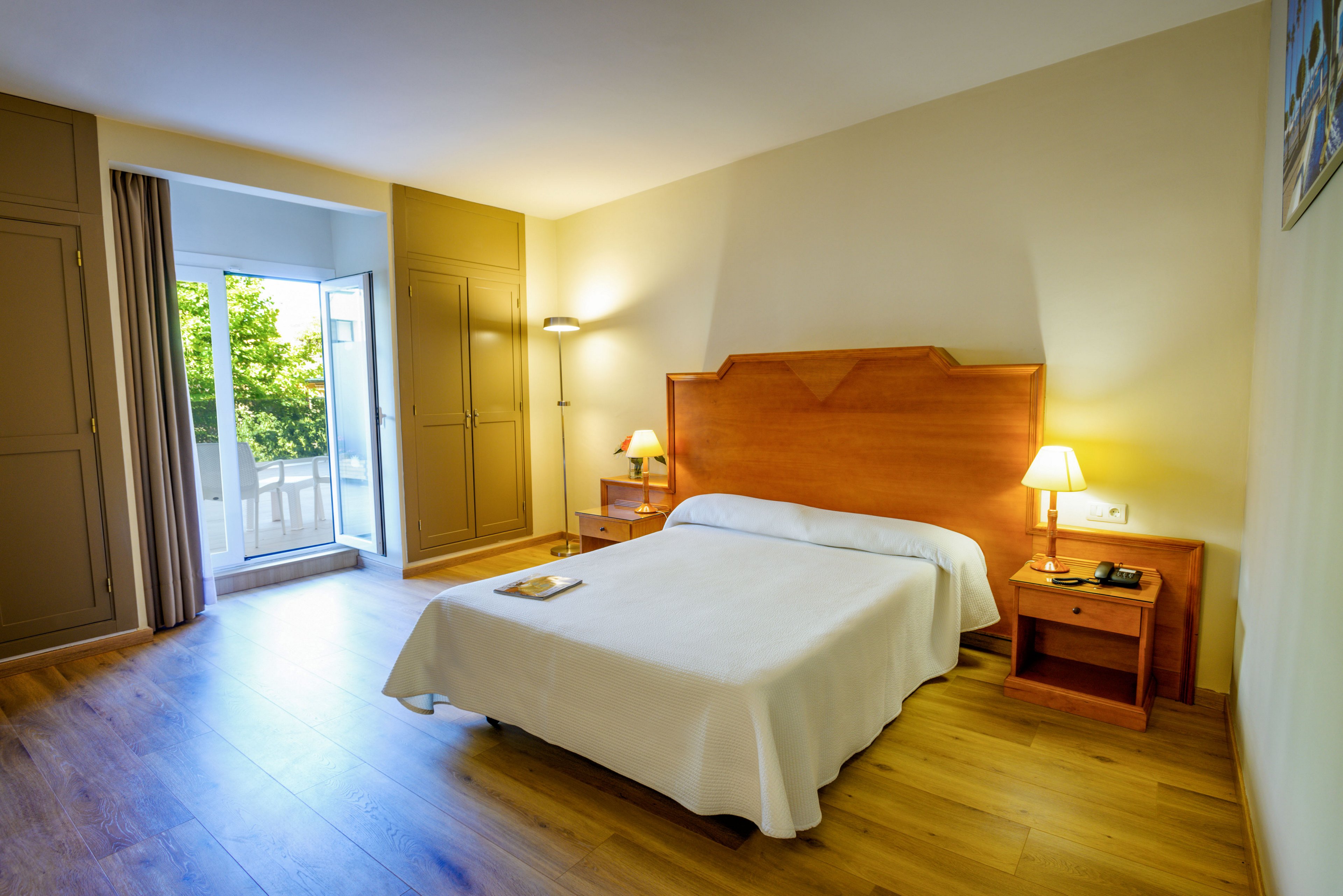 A neatly made double bed with a white bedspread sits centered against a wooden headboard in a hotel-style bedroom. Warm bedside lamps illuminate matching wooden nightstands and a hardwood floor. Built-in wardrobes line the wall, and an open glass door leads to a balcony with greenery visible outside. The overall atmosphere is clean, inviting, and comfortable, suitable for guests seeking a relaxing stay.