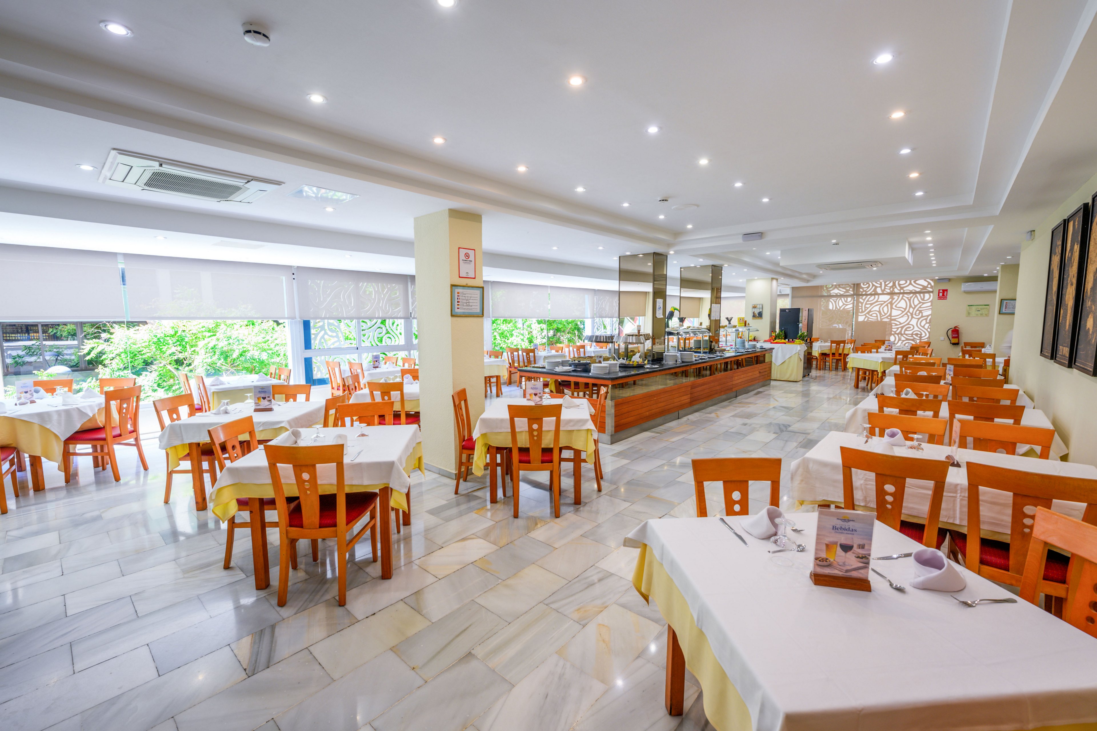 Spacious restaurant dining room with neatly arranged wooden chairs and tables covered in white and yellow tablecloths. The room is empty of people, showcasing a clean and orderly setup with a central buffet area and serving counters. Large windows with blinds let in natural light and reveal greenery outside, while recessed ceiling lights provide even illumination. The polished tile floor and modern decor create a fresh, welcoming atmosphere suitable for hotel dining or events.