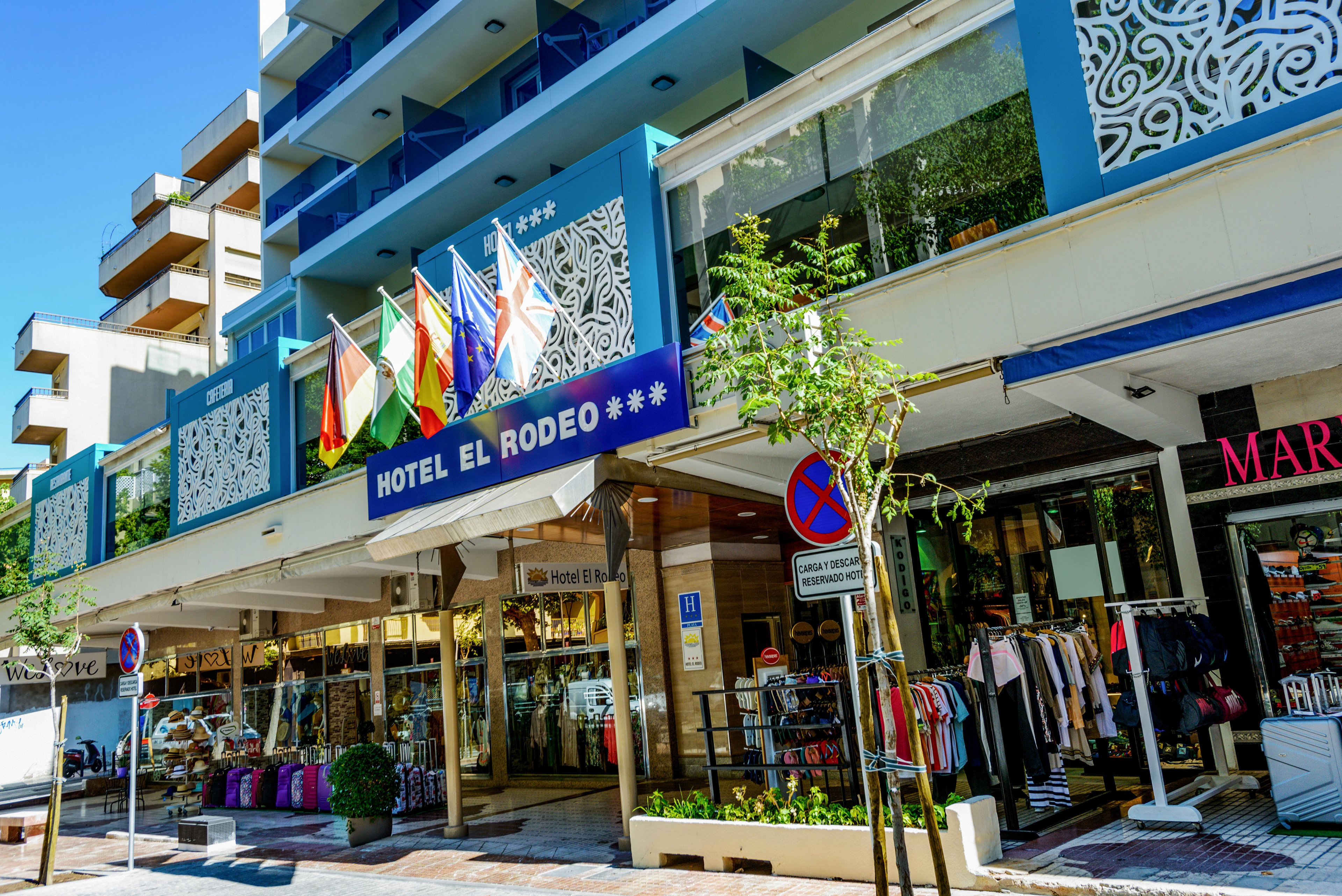 Street-level view of Hotel El Rodeo with its main entrance and blue sign clearly visible. Several international flags are displayed above the doorway, and ground-floor shops line the building with clothing and accessories in the windows. The modern facade features decorative white panels and balconies, suggesting an urban tourist area. A no-parking traffic sign and pedestrian-friendly sidewalk indicate a busy but relaxed city environment.